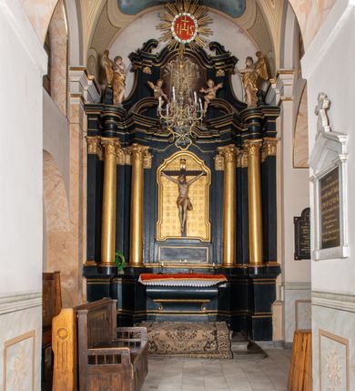 Zdjęcie nr 19: Na dużym krzyżu z ciemnego drewna zawieszono polichromowaną figurę martwego Chrystusa o szeroko rozpostartych ramionach. Ciało Zbawiciela do krzyża jest przymocowane za pomocą trzech gwoździ. On sam został ukazany jako martwy z zamkniętymi oczami i nieznacznie słaniającą się głową. Jego postać jest wysmuklona z realistycznie potraktowaną muskulaturą korpusu. Wyraźnie zaznaczono naprężone ścięgna i mięśnie na całym ciele, co ma sprawiać wrażenie jego autentyczności. Mocno również zaznaczono krwawiącą wybroczynę z przebitego prawego boku. Ciało jest wychudzone o zapadłej klatce piersiowej z silnie podkreśloną linią żeber i mostkiem. Prawa stopa założona jest na lewą. Twarz Zbawiciel jest pociągłą z wyrazistym nosem i wąskim, nieznacznie rozchylonymi ustami. Głowę otaczają brązowe włosy spływające puklami na oba ramiona oraz brązowa broda. Chrystus ma na głowie złoconą, cierniową koronę z długimi cierniami, spod której na czoło spływają krople krwi. Biodra Zbawiciela przepasane są złoconym, krótkim perizonium w formie chusty przerzuconej przez podwójny sznur, odsłaniającej lewe udo i układające się w feston z prawej strony. Ponad głową Chrystusa na pionowej belce krzyża zawieszono rozwiany titulus z napisem &quot;IN / RI&quot;.