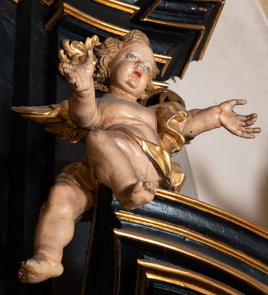 Zdjęcie nr 6: Na dużym krzyżu z ciemnego drewna zawieszono polichromowaną figurę martwego Chrystusa o szeroko rozpostartych ramionach. Ciało Zbawiciela do krzyża jest przymocowane za pomocą trzech gwoździ. On sam został ukazany jako martwy z zamkniętymi oczami i nieznacznie słaniającą się głową. Jego postać jest wysmuklona z realistycznie potraktowaną muskulaturą korpusu. Wyraźnie zaznaczono naprężone ścięgna i mięśnie na całym ciele, co ma sprawiać wrażenie jego autentyczności. Mocno również zaznaczono krwawiącą wybroczynę z przebitego prawego boku. Ciało jest wychudzone o zapadłej klatce piersiowej z silnie podkreśloną linią żeber i mostkiem. Prawa stopa założona jest na lewą. Twarz Zbawiciel jest pociągłą z wyrazistym nosem i wąskim, nieznacznie rozchylonymi ustami. Głowę otaczają brązowe włosy spływające puklami na oba ramiona oraz brązowa broda. Chrystus ma na głowie złoconą, cierniową koronę z długimi cierniami, spod której na czoło spływają krople krwi. Biodra Zbawiciela przepasane są złoconym, krótkim perizonium w formie chusty przerzuconej przez podwójny sznur, odsłaniającej lewe udo i układające się w feston z prawej strony. Ponad głową Chrystusa na pionowej belce krzyża zawieszono rozwiany titulus z napisem &quot;IN / RI&quot;.