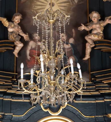 Zdjęcie nr 21: Na dużym krzyżu z ciemnego drewna zawieszono polichromowaną figurę martwego Chrystusa o szeroko rozpostartych ramionach. Ciało Zbawiciela do krzyża jest przymocowane za pomocą trzech gwoździ. On sam został ukazany jako martwy z zamkniętymi oczami i nieznacznie słaniającą się głową. Jego postać jest wysmuklona z realistycznie potraktowaną muskulaturą korpusu. Wyraźnie zaznaczono naprężone ścięgna i mięśnie na całym ciele, co ma sprawiać wrażenie jego autentyczności. Mocno również zaznaczono krwawiącą wybroczynę z przebitego prawego boku. Ciało jest wychudzone o zapadłej klatce piersiowej z silnie podkreśloną linią żeber i mostkiem. Prawa stopa założona jest na lewą. Twarz Zbawiciel jest pociągłą z wyrazistym nosem i wąskim, nieznacznie rozchylonymi ustami. Głowę otaczają brązowe włosy spływające puklami na oba ramiona oraz brązowa broda. Chrystus ma na głowie złoconą, cierniową koronę z długimi cierniami, spod której na czoło spływają krople krwi. Biodra Zbawiciela przepasane są złoconym, krótkim perizonium w formie chusty przerzuconej przez podwójny sznur, odsłaniającej lewe udo i układające się w feston z prawej strony. Ponad głową Chrystusa na pionowej belce krzyża zawieszono rozwiany titulus z napisem "IN / RI".
