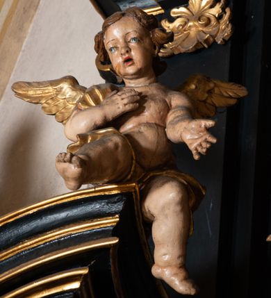 Zdjęcie nr 9: Na dużym krzyżu z ciemnego drewna zawieszono polichromowaną figurę martwego Chrystusa o szeroko rozpostartych ramionach. Ciało Zbawiciela do krzyża jest przymocowane za pomocą trzech gwoździ. On sam został ukazany jako martwy z zamkniętymi oczami i nieznacznie słaniającą się głową. Jego postać jest wysmuklona z realistycznie potraktowaną muskulaturą korpusu. Wyraźnie zaznaczono naprężone ścięgna i mięśnie na całym ciele, co ma sprawiać wrażenie jego autentyczności. Mocno również zaznaczono krwawiącą wybroczynę z przebitego prawego boku. Ciało jest wychudzone o zapadłej klatce piersiowej z silnie podkreśloną linią żeber i mostkiem. Prawa stopa założona jest na lewą. Twarz Zbawiciel jest pociągłą z wyrazistym nosem i wąskim, nieznacznie rozchylonymi ustami. Głowę otaczają brązowe włosy spływające puklami na oba ramiona oraz brązowa broda. Chrystus ma na głowie złoconą, cierniową koronę z długimi cierniami, spod której na czoło spływają krople krwi. Biodra Zbawiciela przepasane są złoconym, krótkim perizonium w formie chusty przerzuconej przez podwójny sznur, odsłaniającej lewe udo i układające się w feston z prawej strony. Ponad głową Chrystusa na pionowej belce krzyża zawieszono rozwiany titulus z napisem "IN / RI".