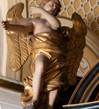 Zdjęcie nr 12: Na dużym krzyżu z ciemnego drewna zawieszono polichromowaną figurę martwego Chrystusa o szeroko rozpostartych ramionach. Ciało Zbawiciela do krzyża jest przymocowane za pomocą trzech gwoździ. On sam został ukazany jako martwy z zamkniętymi oczami i nieznacznie słaniającą się głową. Jego postać jest wysmuklona z realistycznie potraktowaną muskulaturą korpusu. Wyraźnie zaznaczono naprężone ścięgna i mięśnie na całym ciele, co ma sprawiać wrażenie jego autentyczności. Mocno również zaznaczono krwawiącą wybroczynę z przebitego prawego boku. Ciało jest wychudzone o zapadłej klatce piersiowej z silnie podkreśloną linią żeber i mostkiem. Prawa stopa założona jest na lewą. Twarz Zbawiciel jest pociągłą z wyrazistym nosem i wąskim, nieznacznie rozchylonymi ustami. Głowę otaczają brązowe włosy spływające puklami na oba ramiona oraz brązowa broda. Chrystus ma na głowie złoconą, cierniową koronę z długimi cierniami, spod której na czoło spływają krople krwi. Biodra Zbawiciela przepasane są złoconym, krótkim perizonium w formie chusty przerzuconej przez podwójny sznur, odsłaniającej lewe udo i układające się w feston z prawej strony. Ponad głową Chrystusa na pionowej belce krzyża zawieszono rozwiany titulus z napisem "IN / RI".