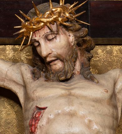 Zdjęcie nr 13: Na dużym krzyżu z ciemnego drewna zawieszono polichromowaną figurę martwego Chrystusa o szeroko rozpostartych ramionach. Ciało Zbawiciela do krzyża jest przymocowane za pomocą trzech gwoździ. On sam został ukazany jako martwy z zamkniętymi oczami i nieznacznie słaniającą się głową. Jego postać jest wysmuklona z realistycznie potraktowaną muskulaturą korpusu. Wyraźnie zaznaczono naprężone ścięgna i mięśnie na całym ciele, co ma sprawiać wrażenie jego autentyczności. Mocno również zaznaczono krwawiącą wybroczynę z przebitego prawego boku. Ciało jest wychudzone o zapadłej klatce piersiowej z silnie podkreśloną linią żeber i mostkiem. Prawa stopa założona jest na lewą. Twarz Zbawiciel jest pociągłą z wyrazistym nosem i wąskim, nieznacznie rozchylonymi ustami. Głowę otaczają brązowe włosy spływające puklami na oba ramiona oraz brązowa broda. Chrystus ma na głowie złoconą, cierniową koronę z długimi cierniami, spod której na czoło spływają krople krwi. Biodra Zbawiciela przepasane są złoconym, krótkim perizonium w formie chusty przerzuconej przez podwójny sznur, odsłaniającej lewe udo i układające się w feston z prawej strony. Ponad głową Chrystusa na pionowej belce krzyża zawieszono rozwiany titulus z napisem "IN / RI".