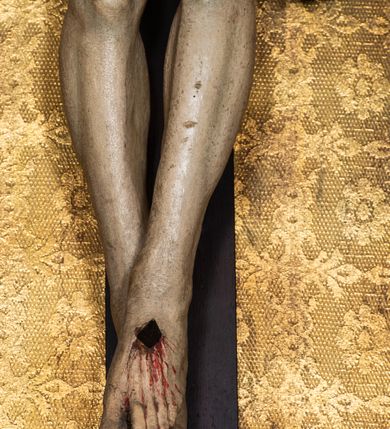 Zdjęcie nr 18: Na dużym krzyżu z ciemnego drewna zawieszono polichromowaną figurę martwego Chrystusa o szeroko rozpostartych ramionach. Ciało Zbawiciela do krzyża jest przymocowane za pomocą trzech gwoździ. On sam został ukazany jako martwy z zamkniętymi oczami i nieznacznie słaniającą się głową. Jego postać jest wysmuklona z realistycznie potraktowaną muskulaturą korpusu. Wyraźnie zaznaczono naprężone ścięgna i mięśnie na całym ciele, co ma sprawiać wrażenie jego autentyczności. Mocno również zaznaczono krwawiącą wybroczynę z przebitego prawego boku. Ciało jest wychudzone o zapadłej klatce piersiowej z silnie podkreśloną linią żeber i mostkiem. Prawa stopa założona jest na lewą. Twarz Zbawiciel jest pociągłą z wyrazistym nosem i wąskim, nieznacznie rozchylonymi ustami. Głowę otaczają brązowe włosy spływające puklami na oba ramiona oraz brązowa broda. Chrystus ma na głowie złoconą, cierniową koronę z długimi cierniami, spod której na czoło spływają krople krwi. Biodra Zbawiciela przepasane są złoconym, krótkim perizonium w formie chusty przerzuconej przez podwójny sznur, odsłaniającej lewe udo i układające się w feston z prawej strony. Ponad głową Chrystusa na pionowej belce krzyża zawieszono rozwiany titulus z napisem "IN / RI".