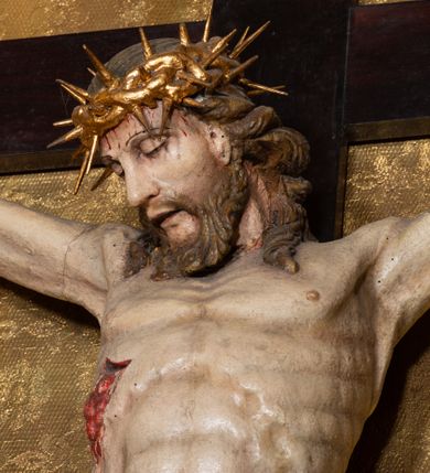 Zdjęcie nr 15: Na dużym krzyżu z ciemnego drewna zawieszono polichromowaną figurę martwego Chrystusa o szeroko rozpostartych ramionach. Ciało Zbawiciela do krzyża jest przymocowane za pomocą trzech gwoździ. On sam został ukazany jako martwy z zamkniętymi oczami i nieznacznie słaniającą się głową. Jego postać jest wysmuklona z realistycznie potraktowaną muskulaturą korpusu. Wyraźnie zaznaczono naprężone ścięgna i mięśnie na całym ciele, co ma sprawiać wrażenie jego autentyczności. Mocno również zaznaczono krwawiącą wybroczynę z przebitego prawego boku. Ciało jest wychudzone o zapadłej klatce piersiowej z silnie podkreśloną linią żeber i mostkiem. Prawa stopa założona jest na lewą. Twarz Zbawiciel jest pociągłą z wyrazistym nosem i wąskim, nieznacznie rozchylonymi ustami. Głowę otaczają brązowe włosy spływające puklami na oba ramiona oraz brązowa broda. Chrystus ma na głowie złoconą, cierniową koronę z długimi cierniami, spod której na czoło spływają krople krwi. Biodra Zbawiciela przepasane są złoconym, krótkim perizonium w formie chusty przerzuconej przez podwójny sznur, odsłaniającej lewe udo i układające się w feston z prawej strony. Ponad głową Chrystusa na pionowej belce krzyża zawieszono rozwiany titulus z napisem "IN / RI".