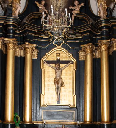 Zdjęcie nr 1: Ołtarz boczny przyjął formę przyściennej, jednokondygnacyjnej i jednoosiowej struktury architektonicznej ze zwieńczeniem. Drewniany stipes ołtarzowy w formie sarkofagu na cokole ustawiono na trójstopniowym podeście. Całą nastawę ustawiono na wysokim, dwustrefowym postumencie, na którym wsparto niski cokół, podtrzymujący zwielokrotnione pilastry o gładkich trzonach i głowicach korynckich oraz flankujący kondygnację dwie pary kolumn. Podpory wspierają wklęsło-wypuklę belkowanie wyłamane na osi kolumn, z umieszczonym na nim na osi pola obrazowego fantazyjnym kartuszem z przedstawieniem gołębicy jako Ducha Świętego. Na belkowaniu, na osi kolumn skrajnych, usytuowano rzeźby stojących aniołów, a na osi kolumn środkowych elementy półkolistego, przerwanego przyczółku z siedzącymi dwoma aniołkami. Zwieńczenie uzyskało kształt leżącego prostokąta o wklęsłych bokach, przechodzących w spływy wolutowe z pilastrami flankującymi pole obrazowe, podtrzymujące wyłamany na osi pilastrów i nadwieszone nad polem obrazowym profilowany gzyms koronujący. Na osi pilastrów ustawiono na nim dwa przyczółki faliste, a na osi retabulum promienistą glorię z chrystogramem "IHS" otoczonym obłokami. Pole obrazowe w kondygnacji uzyskało kształt stojącego prostokąta ujętego profilowaną ramą o nieznacznie wklęsłych bokach i podstawie, zwieńczonego łukiem nadwieszonym z uskokiem. W nim na złoconym tle zawieszono drewniany krucyfiks. Poniżej pola widoczna jest podłużna, prostokątna płycina. W polu zwieńczenia w kształcie obramionego profilem stojącego prostokąta zamkniętego nadwieszonym łukiem odcinkowym zainstalowano obraz z okiem Bożej Opatrzności. Całość retabulum polichromowano na czarno, natomiast kolumny, głowice pilastrów, profile o szaty aniołów i ich skrzydła wyzłocono. Rzeźby aniołów uzyskały polichromię naturalistyczną.    