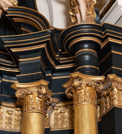 Zdjęcie nr 17: Ołtarz boczny przyjął formę przyściennej, jednokondygnacyjnej i jednoosiowej struktury architektonicznej ze zwieńczeniem. Drewniany stipes ołtarzowy w formie sarkofagu na cokole ustawiono na trójstopniowym podeście. Całą nastawę ustawiono na wysokim, dwustrefowym postumencie, na którym wsparto niski cokół, podtrzymujący zwielokrotnione pilastry o gładkich trzonach i głowicach korynckich oraz flankujący kondygnację dwie pary kolumn. Podpory wspierają wklęsło-wypuklę belkowanie wyłamane na osi kolumn, z umieszczonym na nim na osi pola obrazowego fantazyjnym kartuszem z przedstawieniem gołębicy jako Ducha Świętego. Na belkowaniu, na osi kolumn skrajnych, usytuowano rzeźby stojących aniołów, a na osi kolumn środkowych elementy półkolistego, przerwanego przyczółku z siedzącymi dwoma aniołkami. Zwieńczenie uzyskało kształt leżącego prostokąta o wklęsłych bokach, przechodzących w spływy wolutowe z pilastrami flankującymi pole obrazowe, podtrzymujące wyłamany na osi pilastrów i nadwieszone nad polem obrazowym profilowany gzyms koronujący. Na osi pilastrów ustawiono na nim dwa przyczółki faliste, a na osi retabulum promienistą glorię z chrystogramem "IHS" otoczonym obłokami. Pole obrazowe w kondygnacji uzyskało kształt stojącego prostokąta ujętego profilowaną ramą o nieznacznie wklęsłych bokach i podstawie, zwieńczonego łukiem nadwieszonym z uskokiem. W nim na złoconym tle zawieszono drewniany krucyfiks. Poniżej pola widoczna jest podłużna, prostokątna płycina. W polu zwieńczenia w kształcie obramionego profilem stojącego prostokąta zamkniętego nadwieszonym łukiem odcinkowym zainstalowano obraz z okiem Bożej Opatrzności. Całość retabulum polichromowano na czarno, natomiast kolumny, głowice pilastrów, profile o szaty aniołów i ich skrzydła wyzłocono. Rzeźby aniołów uzyskały polichromię naturalistyczną.    