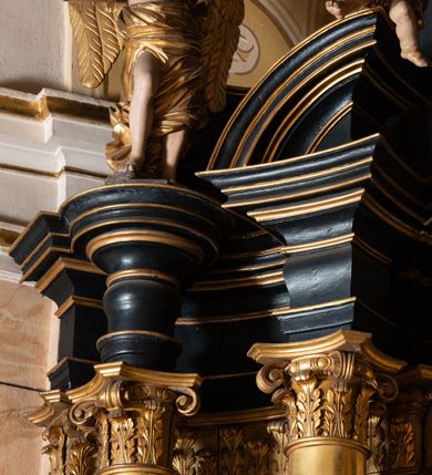 Zdjęcie nr 18: Ołtarz boczny przyjął formę przyściennej, jednokondygnacyjnej i jednoosiowej struktury architektonicznej ze zwieńczeniem. Drewniany stipes ołtarzowy w formie sarkofagu na cokole ustawiono na trójstopniowym podeście. Całą nastawę ustawiono na wysokim, dwustrefowym postumencie, na którym wsparto niski cokół, podtrzymujący zwielokrotnione pilastry o gładkich trzonach i głowicach korynckich oraz flankujący kondygnację dwie pary kolumn. Podpory wspierają wklęsło-wypuklę belkowanie wyłamane na osi kolumn, z umieszczonym na nim na osi pola obrazowego fantazyjnym kartuszem z przedstawieniem gołębicy jako Ducha Świętego. Na belkowaniu, na osi kolumn skrajnych, usytuowano rzeźby stojących aniołów, a na osi kolumn środkowych elementy półkolistego, przerwanego przyczółku z siedzącymi dwoma aniołkami. Zwieńczenie uzyskało kształt leżącego prostokąta o wklęsłych bokach, przechodzących w spływy wolutowe z pilastrami flankującymi pole obrazowe, podtrzymujące wyłamany na osi pilastrów i nadwieszone nad polem obrazowym profilowany gzyms koronujący. Na osi pilastrów ustawiono na nim dwa przyczółki faliste, a na osi retabulum promienistą glorię z chrystogramem "IHS" otoczonym obłokami. Pole obrazowe w kondygnacji uzyskało kształt stojącego prostokąta ujętego profilowaną ramą o nieznacznie wklęsłych bokach i podstawie, zwieńczonego łukiem nadwieszonym z uskokiem. W nim na złoconym tle zawieszono drewniany krucyfiks. Poniżej pola widoczna jest podłużna, prostokątna płycina. W polu zwieńczenia w kształcie obramionego profilem stojącego prostokąta zamkniętego nadwieszonym łukiem odcinkowym zainstalowano obraz z okiem Bożej Opatrzności. Całość retabulum polichromowano na czarno, natomiast kolumny, głowice pilastrów, profile o szaty aniołów i ich skrzydła wyzłocono. Rzeźby aniołów uzyskały polichromię naturalistyczną.    
