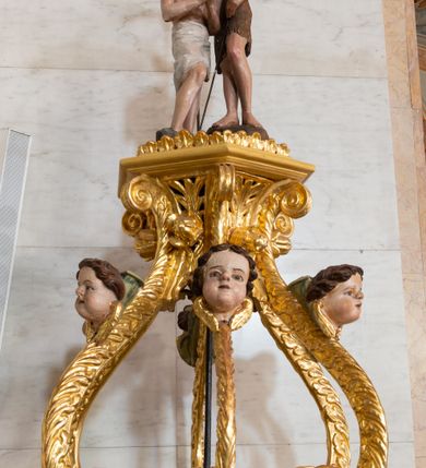 Zdjęcie nr 3: Chrzcielnica uzyskała kształt spiętrzonych jeden na drugim trzech drewnianych prostopadłościanów na rzucie foremnego sześcioboku ustawionych diagonalnie w stosunku do czwartego modułu, jakim jest sześcioboczny cokół także wykonany z drewna. Najniższy prostopadłościan od cokołu oddzielony jest profilowaną listwą, powtórzoną na jego zwieńczeniu w formie gzymsu, będącego jednocześnie cokolikiem dla trzeciego prostopadłościanu zamkniętego od góry profilowanym belkowaniem, na którym wspiera się dekoracyjna przykrywa. Boki pierwszego poziomu są gładkie, natomiast środkowego i ostatniego zostały ozdobione na krawędziach spływami wolutowymi oraz podwieszonym u góry ornamentem z motywem stylizowanych listków. Pomiędzy wolutami środkowego poziomu na bokach prostopadłościanu umieszczono uskrzydlone główki anielskie, natomiast górnego kwiat róży ujęty przez dwa listki. Przykrywę tworzy sześć ażurowo ustawionych esowato wygiętych wolut roślinnych przechodzących u góry w stylizowany kapitel kompozytowy. Na nim umieszczono sześcioboczny, profilowany niski cokolik zwieńczony sceną chrztu w Jordanie. Woluty przykrywy wokół kapitelu ozdobione zostały rzeźbionymi, uskrzydlonymi główkami aniołków. Na szczycie stoi w lekkim zgięciu Chrystus ze złożonymi przed sobą modlitewnie rękami, który przyjmuje chrzest od stojącego obok Jana Chrzciciela. Ponad nimi unosi się gołębica Ducha Świętego. 
