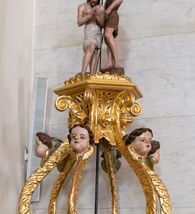 Zdjęcie nr 2: Chrzcielnica uzyskała kształt spiętrzonych jeden na drugim trzech drewnianych prostopadłościanów na rzucie foremnego sześcioboku ustawionych diagonalnie w stosunku do czwartego modułu, jakim jest sześcioboczny cokół także wykonany z drewna. Najniższy prostopadłościan od cokołu oddzielony jest profilowaną listwą, powtórzoną na jego zwieńczeniu w formie gzymsu, będącego jednocześnie cokolikiem dla trzeciego prostopadłościanu zamkniętego od góry profilowanym belkowaniem, na którym wspiera się dekoracyjna przykrywa. Boki pierwszego poziomu są gładkie, natomiast środkowego i ostatniego zostały ozdobione na krawędziach spływami wolutowymi oraz podwieszonym u góry ornamentem z motywem stylizowanych listków. Pomiędzy wolutami środkowego poziomu na bokach prostopadłościanu umieszczono uskrzydlone główki anielskie, natomiast górnego kwiat róży ujęty przez dwa listki. Przykrywę tworzy sześć ażurowo ustawionych esowato wygiętych wolut roślinnych przechodzących u góry w stylizowany kapitel kompozytowy. Na nim umieszczono sześcioboczny, profilowany niski cokolik zwieńczony sceną chrztu w Jordanie. Woluty przykrywy wokół kapitelu ozdobione zostały rzeźbionymi, uskrzydlonymi główkami aniołków. Na szczycie stoi w lekkim zgięciu Chrystus ze złożonymi przed sobą modlitewnie rękami, który przyjmuje chrzest od stojącego obok Jana Chrzciciela. Ponad nimi unosi się gołębica Ducha Świętego. 