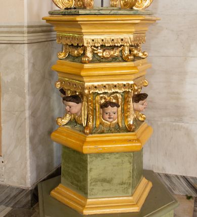 Zdjęcie nr 1: Chrzcielnica uzyskała kształt spiętrzonych jeden na drugim trzech drewnianych prostopadłościanów na rzucie foremnego sześcioboku ustawionych diagonalnie w stosunku do czwartego modułu, jakim jest sześcioboczny cokół także wykonany z drewna. Najniższy prostopadłościan od cokołu oddzielony jest profilowaną listwą, powtórzoną na jego zwieńczeniu w formie gzymsu, będącego jednocześnie cokolikiem dla trzeciego prostopadłościanu zamkniętego od góry profilowanym belkowaniem, na którym wspiera się dekoracyjna przykrywa. Boki pierwszego poziomu są gładkie, natomiast środkowego i ostatniego zostały ozdobione na krawędziach spływami wolutowymi oraz podwieszonym u góry ornamentem z motywem stylizowanych listków. Pomiędzy wolutami środkowego poziomu na bokach prostopadłościanu umieszczono uskrzydlone główki anielskie, natomiast górnego kwiat róży ujęty przez dwa listki. Przykrywę tworzy sześć ażurowo ustawionych esowato wygiętych wolut roślinnych przechodzących u góry w stylizowany kapitel kompozytowy. Na nim umieszczono sześcioboczny, profilowany niski cokolik zwieńczony sceną chrztu w Jordanie. Woluty przykrywy wokół kapitelu ozdobione zostały rzeźbionymi, uskrzydlonymi główkami aniołków. Na szczycie stoi w lekkim zgięciu Chrystus ze złożonymi przed sobą modlitewnie rękami, który przyjmuje chrzest od stojącego obok Jana Chrzciciela. Ponad nimi unosi się gołębica Ducha Świętego. 