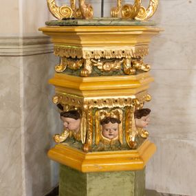 Zdjęcie nr 1: Chrzcielnica uzyskała kształt spiętrzonych jeden na drugim trzech drewnianych prostopadłościanów na rzucie foremnego sześcioboku ustawionych diagonalnie w stosunku do czwartego modułu, jakim jest sześcioboczny cokół także wykonany z drewna. Najniższy prostopadłościan od cokołu oddzielony jest profilowaną listwą, powtórzoną na jego zwieńczeniu w formie gzymsu, będącego jednocześnie cokolikiem dla trzeciego prostopadłościanu zamkniętego od góry profilowanym belkowaniem, na którym wspiera się dekoracyjna przykrywa. Boki pierwszego poziomu są gładkie, natomiast środkowego i ostatniego zostały ozdobione na krawędziach spływami wolutowymi oraz podwieszonym u góry ornamentem z motywem stylizowanych listków. Pomiędzy wolutami środkowego poziomu na bokach prostopadłościanu umieszczono uskrzydlone główki anielskie, natomiast górnego kwiat róży ujęty przez dwa listki. Przykrywę tworzy sześć ażurowo ustawionych esowato wygiętych wolut roślinnych przechodzących u góry w stylizowany kapitel kompozytowy. Na nim umieszczono sześcioboczny, profilowany niski cokolik zwieńczony sceną chrztu w Jordanie. Woluty przykrywy wokół kapitelu ozdobione zostały rzeźbionymi, uskrzydlonymi główkami aniołków. Na szczycie stoi w lekkim zgięciu Chrystus ze złożonymi przed sobą modlitewnie rękami, który przyjmuje chrzest od stojącego obok Jana Chrzciciela. Ponad nimi unosi się gołębica Ducha Świętego. 