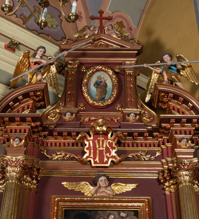 Zdjęcie nr 4: Ołtarz architektoniczny, jednoosiowy, jednokondygnacyjny ze zwieńczeniem. Retabulum osadzone jest na wysokim, zdwojonym, zryzalitowanym uskokowo cokole. Ołtarz jest prostopadłościenny z drewnianą mensą. Część środkową ujmuje para kolumn o żłobkowanych trzonach i korynckich kapitelach, ustawionych na tle zdwojonych pilastrów. Podpory dźwigają wyłamujące się ponad nimi belkowanie o silnie wysuniętym gzymsie. Zwieńczenie ma formę aediculi ujętej parą pilastrów i spływów wolutowych, zamkniętej trójkątnym przyczółkiem. Dodatkowo ujęta jest ona przerwanym, półkolistym przyczółkiem. Płycina antepedium jest w kształcie leżącego prostokąta z płaskorzeźbionym motywem złoconego kielicha eucharystycznego oraz kwiatów lilii skrzyżowanych z włócznią oraz trzciną z gąbką na tle srebrzonych obłoków ujętych promienistą glorią. Płyciny górnego cokołu (predelli) zdobi motyw ostów i cierni. Część środkową ujmuje para ażurowych, akantowych uszaków z motywem tuniki, kolumny biczowania oraz trzciny po lewej stronie i chusty św. Weroniki, drabiny, włóczni i trzciny z gąbką po prawej. U góry, pośrodku znajduje się uskrzydlona główka anielska. Powyżej, w partii belkowania, którego gzyms zdobi kostkowanie, jest umieszczony kartusz herbowy z monogramem IHS. Fryz zdobi stylizowana wić roślinna oraz para uskrzydlonych główek anielskich, natomiast zwieńczenie retabulum m.in. motywy roślinne, rozetki i kampanulle. Szczyt tworzy podtrzymująca krzyż para aniołów o sylwetkach od pasa w dół w formie liści akantu. Na przerwanym przyczółku znajduje się para przyklękających aniołów, ubranych w suknie w kolorach jasnoczerwonym i niebieskim oraz złocone płaszcze, trzymających w dłoniach włócznię i trzcinę z gąbką. Pole w części środkowej ma kształt stojącego prostokąta ujętego profilowaną, złoconą ramą. W środku znajduje się przedstawienie św. Anny nauczającej małą Marię. Pole w zwieńczeniu ma kształt stojącego owalu ujętego złoconą ramą w formie wieńca laurowego. Zawiera wizerunek św. Józefa. 
Struktura retabulum utrzymana jest w odcieniach brązu, partie detalu architektonicznego i ornamentyka są złocone.      
