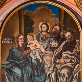 Zdjęcie nr 1: Obraz w kształcie zbliżonym do kwadratu zamkniętego łukiem półkolistym przedstawia wielopostaciową kompozycję zatytułowaną Święta Rodzina. Pośrodku, na podwyższeniu ukazana jest siedząca postać Matki Boskiej zwróconej na wprost i kierującej głowę w trzech czwartych w prawo. Owalną twarz charakteryzują młodzieńcze rysy. Maria ubrana jest w białą i nałożoną na nią jasnobrązową suknię oraz niebieski płaszcz częściowo osłaniający zwieńczoną koroną głowę. Na kolanach trzyma, osłaniając je białą chustą, półnagie Dzieciątko przechylające się w prawo, z koroną na głowie. Jezus wyciąga rączki w kierunku siedzącej obok św. Anny. Zwrócona w trzech czwartych w lewo kobieta ubrana jest w brązową i ciemnoniebieską suknię oraz ciemnoniebieski płaszcz. Głowę osłania brązowa chusta. Twarz Anny charakteryzują wyraziste rysy. Święta oburącz ujmuje Jezusa. W tle, po lewej stronie za Marią widoczna jest postać św. Józefa zwróconego w trzech czwartych w prawo, trzymającego kwiat białej lilii. Nieco bliżej siedzi zwrócona w trzech czwartych w prawo sylwetka św. Joachima. Jego twarz charakteryzują rysy mężczyzny w podeszłym wieku oraz siwe zarost i włosy. Święty ubrany jest w bordową suknię i popielaty płaszcz. Lewą rękę unosi na wysokości klatki piersiowej, prawą podtrzymuje leżącą na kolanach otwartą księgę. Głowę Marii i Jezusa okalają świetliste nimby, pozostałych postaci aureole. Scena ukazana jest w pomieszczeniu otwierającym się na zewnątrz dużym oknem. Na pierwszym planie, po bokach znajduje się para wazonów z kwiatami. Po prawej stronie obrazu, na wysokości głowy św. Anny jest napis „SACRA / FAMILIA”.           