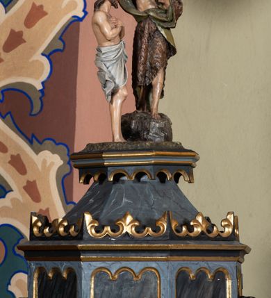Zdjęcie nr 2: Chrzcielnica na rzucie ośmioboku, trzon i czara również są ośmioboczne. Cokół dwustopniowy, stopnie oddzielone od siebie zaoblonym uskokiem, w górnej części łagodnie przechodzą w trzon. W środkowej partii trzonu spłaszczony ośmioboczny pierścień dekorowany taśmą z płaskorzeźbionymi rozetkami. Czara rozchylająca się ku górze, z profilowaną podstawą, rozczłonkowana ostrołukowatymi płycinami, zakończona profilowanym gzymsem. Ośmioboczna pokrywa o polach z wyciętymi płycinami zamkniętymi potrójnymi łukami półkolistymi jest zwieńczona profilowanym gzymsem z koronką i nakryta daszkiem. Nad nim podstawa ujęta od dołu arkadkowaniem z grupą rzeźbiarską Chrztu w Jordanie. Chrystus stoi w rzece z rękami założonymi na piersi, opasany na biodrach długą białą tkaniną. Przed nim, na skale stoi św. Jan Chrzciciel, ubrany w wielbłądzią skórę z prawą ręką trzymającą naczynie z wodą nad głową Chrystusa, a lewą wyciągniętą do przodu. Nad nim unosi się gołębica Ducha Świętego. 