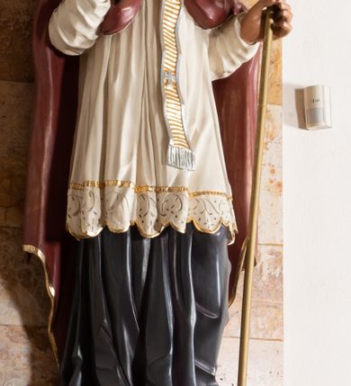 Zdjęcie nr 3: Rzeźba drążona z tyłu stoi na prostokątnej podstawie ustawionej na profilowanej, zwężającej się ku dołowi konsoli przyściennej. Postać biskupa jest ujęta frontalni w nieznacznym kontrapoście z głową lekko skierowaną w prawo. Prawą rękę Augustyn ma uniesioną przed siebie na wysokość piersi w lewej zaś trzyma pastorał o gładkiej, zamkniętej krzywaśni z wpisanym krzyżem równoramiennym. Święty ma pociągłą, wychudzoną twarz o szeroko otwartych oczach, mięsistym nosie i pełnych ustach. Głowę okala broda i lekko kręcone sięgające ramion włosy. Augustyn na głowie ma szkarłatną mitrę zdobiną złotą ornamentyką. Ubrany jest w sutannę, białą komże dekorowaną u spodu szeroką koronką oraz szkarłatną kapę pokrytą złocistym ornamentem i lamowaną złotą tasiemką. Na ramionach dodatkowo ma założony złocony i srebrzony paliusz. Na postumencie widoczny jest podpis: "Ś[WIĘTY] AUGUSTYN".