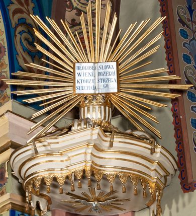 Zdjęcie nr 5: Ambona przyścienna, wisząca, z dostępem przez drzwi w partii zaplecka. Kosz na planie zbliżonym do koła o wklęsło-wypukłym zarysie, na cokole w postaci spłaszczonego wałka, z podwieszeniem w formie zbliżonej do spłaszczonej półkuli, zdobionej rozetami, ujętej lizenami dekorowanymi zwisami, zakończonej szyszką. Ścianki kosza wydzielone pilastrami dźwigającymi belkowanie tworzące parapet. Trzony dekorowane zwisami, ścianki rozetami. Na krawędzi cokołu siedzące figury czterech ewangelistów. Baldachim na planie analogicznym do kosza w formie belkowania z podwieszonym lambrekinem. Na nim woluty tworzące cokół dla wazonu, na którym znajduje się otwarta księga w glorii promienistej. Na księdze napis: „BŁOGOSŁA/WIENI / KTÓRZY / SŁUCHAIĄ / SŁOWA / BOŻEGO, I / STRZEGĄ / GO. / U S(więtego) Łukasza w rozdz(iale) XI wier(sz) [28]”. Na podniebieniu baldachimu gołębica Ducha Świętego w glorii promienistej. Struktura polichromowana w kolorze beżowym, ze złoconymi profilami, ornamentyką i detalami. 