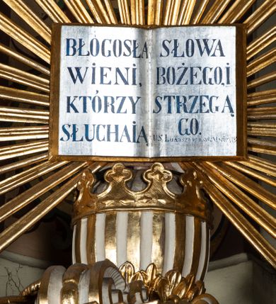 Zdjęcie nr 6: Ambona przyścienna, wisząca, z dostępem przez drzwi w partii zaplecka. Kosz na planie zbliżonym do koła o wklęsło-wypukłym zarysie, na cokole w postaci spłaszczonego wałka, z podwieszeniem w formie zbliżonej do spłaszczonej półkuli, zdobionej rozetami, ujętej lizenami dekorowanymi zwisami, zakończonej szyszką. Ścianki kosza wydzielone pilastrami dźwigającymi belkowanie tworzące parapet. Trzony dekorowane zwisami, ścianki rozetami. Na krawędzi cokołu siedzące figury czterech ewangelistów. Baldachim na planie analogicznym do kosza w formie belkowania z podwieszonym lambrekinem. Na nim woluty tworzące cokół dla wazonu, na którym znajduje się otwarta księga w glorii promienistej. Na księdze napis: „BŁOGOSŁA/WIENI / KTÓRZY / SŁUCHAIĄ / SŁOWA / BOŻEGO, I / STRZEGĄ / GO. / U S(więtego) Łukasza w rozdz(iale) XI wier(sz) [28]”. Na podniebieniu baldachimu gołębica Ducha Świętego w glorii promienistej. Struktura polichromowana w kolorze beżowym, ze złoconymi profilami, ornamentyką i detalami. 