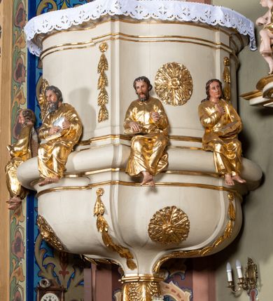 Zdjęcie nr 4: Ambona przyścienna, wisząca, z dostępem przez drzwi w partii zaplecka. Kosz na planie zbliżonym do koła o wklęsło-wypukłym zarysie, na cokole w postaci spłaszczonego wałka, z podwieszeniem w formie zbliżonej do spłaszczonej półkuli, zdobionej rozetami, ujętej lizenami dekorowanymi zwisami, zakończonej szyszką. Ścianki kosza wydzielone pilastrami dźwigającymi belkowanie tworzące parapet. Trzony dekorowane zwisami, ścianki rozetami. Na krawędzi cokołu siedzące figury czterech ewangelistów. Baldachim na planie analogicznym do kosza w formie belkowania z podwieszonym lambrekinem. Na nim woluty tworzące cokół dla wazonu, na którym znajduje się otwarta księga w glorii promienistej. Na księdze napis: „BŁOGOSŁA/WIENI / KTÓRZY / SŁUCHAIĄ / SŁOWA / BOŻEGO, I / STRZEGĄ / GO. / U S(więtego) Łukasza w rozdz(iale) XI wier(sz) [28]”. Na podniebieniu baldachimu gołębica Ducha Świętego w glorii promienistej. Struktura polichromowana w kolorze beżowym, ze złoconymi profilami, ornamentyką i detalami. 