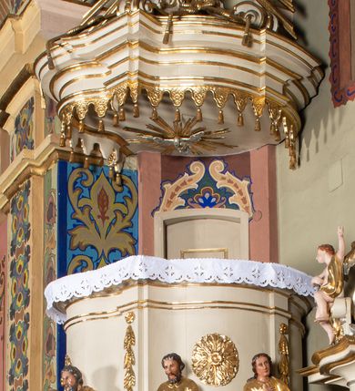Zdjęcie nr 2: Ambona przyścienna, wisząca, z dostępem przez drzwi w partii zaplecka. Kosz na planie zbliżonym do koła o wklęsło-wypukłym zarysie, na cokole w postaci spłaszczonego wałka, z podwieszeniem w formie zbliżonej do spłaszczonej półkuli, zdobionej rozetami, ujętej lizenami dekorowanymi zwisami, zakończonej szyszką. Ścianki kosza wydzielone pilastrami dźwigającymi belkowanie tworzące parapet. Trzony dekorowane zwisami, ścianki rozetami. Na krawędzi cokołu siedzące figury czterech ewangelistów. Baldachim na planie analogicznym do kosza w formie belkowania z podwieszonym lambrekinem. Na nim woluty tworzące cokół dla wazonu, na którym znajduje się otwarta księga w glorii promienistej. Na księdze napis: „BŁOGOSŁA/WIENI / KTÓRZY / SŁUCHAIĄ / SŁOWA / BOŻEGO, I / STRZEGĄ / GO. / U S(więtego) Łukasza w rozdz(iale) XI wier(sz) [28]”. Na podniebieniu baldachimu gołębica Ducha Świętego w glorii promienistej. Struktura polichromowana w kolorze beżowym, ze złoconymi profilami, ornamentyką i detalami. 