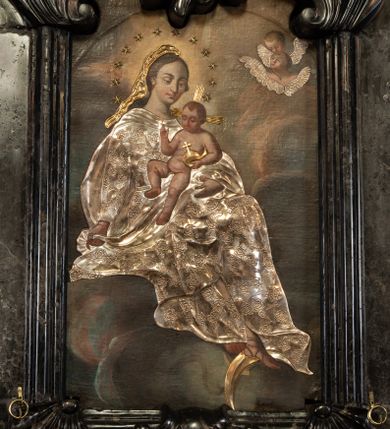 Zdjęcie nr 1: Obraz w formie stojącego prostokąta. W centrum na obłokach siedzi Maria, zwrócona w trzech czwartych w lewo, lewą ręką podtrzymuje Dzieciątko, w prawej, wyciągniętej w bok trzyma fragment płaszcza. Stopy opiera na sierpie księżyca, przesłoniętym metalowym, wokół którego wije się wąż. Maria ma owalną twarz, z wysokim czołem, o dużych, okrągłych oczach skierowanych w dół, dużym nosie i małych, pełnych ustach; okoloną długimi, brązowymi włosami. Wokół głowy ma wieniec z aplikowanych, metalowych gwiazd. Ubrana jest w strój przesłonięty metalową, srebrzoną sukienką w postaci sukni o kwiatowym wzorze, przepasaną w talii, na plecy ma narzucony płaszcz, zawinięty na jej lewym ramieniu, na którym siedzi Dzieciątko. Na głowie ma welon przesłonięty metalowym, złoconym. Jezus w pozycji siedzącej, zwrócony w trzech czwartych w prawo, w lewej dłoni trzyma jabłko królewskie, przesłonięte metalowym, złoconym, prawą unosi w geście błogosławieństwa. Twarz ma owalną, o okrągłych oczach skierowanych w dół, małym nosie i pełnych ustach, okoloną krótkimi, jasnobrązowymi włosami. Ciało ma nagie, pulchne. Od jego głowy odchodzą trzy wiązki aplikowanych, metalowych i złoconych promieni. Postacie ukazane są na tle gęstych, szaro-różowych obłoków, rozświetlonych na żółto w górnej części obrazu, wokół głowy Marii. W prawym górnym rogu dwie uskrzydlone główki anielskie z aplikowanmi metalowymi i srebrzonymi skrzydłami. 