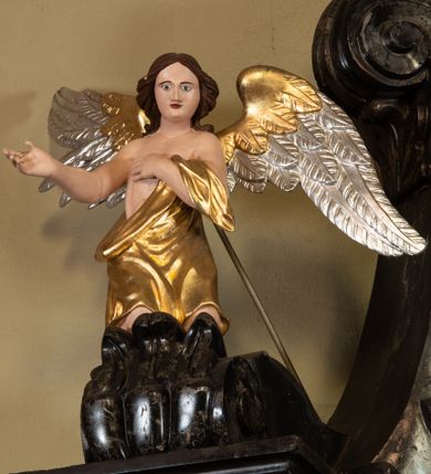 Zdjęcie nr 1: Anioł ukazany frontalnie, w pozycji klęczącej, lewą rękę kładzie na piersi, prawą wyciąga w bok. Twarz ma okrągłą, o dużych, migdałowatych oczach, prostym nosie i małych ustach; okoloną brązowymi, kręconymi włosami sięgającymi ramion. Ubrany jest w złocony płaszcz, przesłaniający go w partii nóg, odsłaniający tors, zarzucony na lewe przedramię. Skrzydła ma duże, rozłożone, częściowo złocone i srebrzone. Polichromia ciała naturalistyczna. 