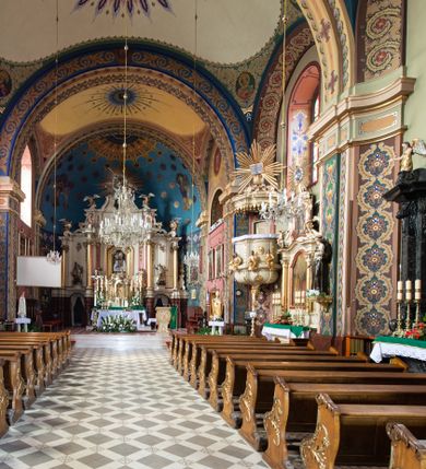 Zdjęcie nr 5: Kościół murowany, tynkowany, na kamiennym podmurowaniu, orientowany, jednonawowy. Prezbiterium prostokątne, krótkie, zamknięte trójbocznie, przy południowej ścianie prostokątna zakrystia z piętrem, z prostokątnym aneksem od zachodu. Nawa główna szersza od prezbiterium, na rzucie prostokąta, od zachodu przylega prostokątny przedsionek z wieżą na planie kwadratu. Wnętrze podzielone na cztery prostokątne przęsła i absydę, wydzielone arkadami, z gurtami spływającymi na zwielokrotnione pilastry. Każde przęsło nakryte sklepieniem o zaokrąglonych narożach. Łuk tęczowy półkolisty. W zachodniej części parapet chóru muzycznego wsparty na dwóch filarach i dwóch półfilarach, tworzących trzy arkady. Fasada trójosiowa, z wbudowaną wieżą, ujętą spłaszczonymi spływami zakończonymi cokołami z figurami. Osie wydzielone lizenami, podtrzymującymi profilowany gzyms, pod nim malowany fryz z dekoracją roślinną. Na osi portal w formie stojącego prostokąta zamkniętego łukiem odcinkowym, ujęty opaską tynkową, zakończoną kluczem. Drzwi drewniane, dwuskrzydłowe. Nad portalem fragment profilowanego gzymsu z daszkiem okapowym. Powyżej, na osi okno w formie stojącego prostokąta zamkniętego łukiem odcinkowym, w profilowanej opasce tynkowej, ujętej malowaną dekoracją geometryczną. Wieża dwukondygnacyjna, flankowana w narożach lizenami podtrzymującymi profilowany gzyms wieńczący. Na ścianie frontowej dwa okna na każdej kondygnacji, w formie stojącego prostokąta zamkniętego łukiem odcinkowym, w profilowanej opasce tynkowej, otoczonej malowaną dekoracją geometryczną, zwieńczone tarczą zegarową. Na pozostałych trzech ścianach wieży po jednym analogicznym oknie w drugiej kondygnacji i tarczy zegarowej. Elewacje północna i południowa artykułowane lizenami podtrzymującymi profilowany gzyms, z dekoracyjnym fryzem poniżej. Okna w formie stojącego prostokąta zamkniętego łukiem odcinkowym, w profilowanej opasce tynkowej z kluczem, ujętej malowaną dekoracją geometryczną: cztery w północnej i trzy w południowej. W zakrystii jedno prostokątne okno od południa i dwa w każdej kondygnacji od wschodu. Nawa główna i prezbiterium nakryte osobnymi dachami dwuspadowymi. Na ich połączeniu wieżyczka na sygnaturkę z latarnią, zwieńczona hełmem baniastym z krzyżem. Wieża nakryta hełmem baniastym zakończonym kulą z krzyżem. Zakrystia przykryta dachem trójspadowym, a aneks czterospadowym. 