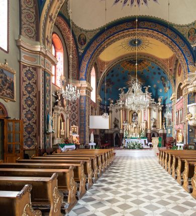 Zdjęcie nr 4: Kościół murowany, tynkowany, na kamiennym podmurowaniu, orientowany, jednonawowy. Prezbiterium prostokątne, krótkie, zamknięte trójbocznie, przy południowej ścianie prostokątna zakrystia z piętrem, z prostokątnym aneksem od zachodu. Nawa główna szersza od prezbiterium, na rzucie prostokąta, od zachodu przylega prostokątny przedsionek z wieżą na planie kwadratu. Wnętrze podzielone na cztery prostokątne przęsła i absydę, wydzielone arkadami, z gurtami spływającymi na zwielokrotnione pilastry. Każde przęsło nakryte sklepieniem o zaokrąglonych narożach. Łuk tęczowy półkolisty. W zachodniej części parapet chóru muzycznego wsparty na dwóch filarach i dwóch półfilarach, tworzących trzy arkady. Fasada trójosiowa, z wbudowaną wieżą, ujętą spłaszczonymi spływami zakończonymi cokołami z figurami. Osie wydzielone lizenami, podtrzymującymi profilowany gzyms, pod nim malowany fryz z dekoracją roślinną. Na osi portal w formie stojącego prostokąta zamkniętego łukiem odcinkowym, ujęty opaską tynkową, zakończoną kluczem. Drzwi drewniane, dwuskrzydłowe. Nad portalem fragment profilowanego gzymsu z daszkiem okapowym. Powyżej, na osi okno w formie stojącego prostokąta zamkniętego łukiem odcinkowym, w profilowanej opasce tynkowej, ujętej malowaną dekoracją geometryczną. Wieża dwukondygnacyjna, flankowana w narożach lizenami podtrzymującymi profilowany gzyms wieńczący. Na ścianie frontowej dwa okna na każdej kondygnacji, w formie stojącego prostokąta zamkniętego łukiem odcinkowym, w profilowanej opasce tynkowej, otoczonej malowaną dekoracją geometryczną, zwieńczone tarczą zegarową. Na pozostałych trzech ścianach wieży po jednym analogicznym oknie w drugiej kondygnacji i tarczy zegarowej. Elewacje północna i południowa artykułowane lizenami podtrzymującymi profilowany gzyms, z dekoracyjnym fryzem poniżej. Okna w formie stojącego prostokąta zamkniętego łukiem odcinkowym, w profilowanej opasce tynkowej z kluczem, ujętej malowaną dekoracją geometryczną: cztery w północnej i trzy w południowej. W zakrystii jedno prostokątne okno od południa i dwa w każdej kondygnacji od wschodu. Nawa główna i prezbiterium nakryte osobnymi dachami dwuspadowymi. Na ich połączeniu wieżyczka na sygnaturkę z latarnią, zwieńczona hełmem baniastym z krzyżem. Wieża nakryta hełmem baniastym zakończonym kulą z krzyżem. Zakrystia przykryta dachem trójspadowym, a aneks czterospadowym. 