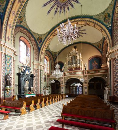 Zdjęcie nr 10: Kościół murowany, tynkowany, na kamiennym podmurowaniu, orientowany, jednonawowy. Prezbiterium prostokątne, krótkie, zamknięte trójbocznie, przy południowej ścianie prostokątna zakrystia z piętrem, z prostokątnym aneksem od zachodu. Nawa główna szersza od prezbiterium, na rzucie prostokąta, od zachodu przylega prostokątny przedsionek z wieżą na planie kwadratu. Wnętrze podzielone na cztery prostokątne przęsła i absydę, wydzielone arkadami, z gurtami spływającymi na zwielokrotnione pilastry. Każde przęsło nakryte sklepieniem o zaokrąglonych narożach. Łuk tęczowy półkolisty. W zachodniej części parapet chóru muzycznego wsparty na dwóch filarach i dwóch półfilarach, tworzących trzy arkady. Fasada trójosiowa, z wbudowaną wieżą, ujętą spłaszczonymi spływami zakończonymi cokołami z figurami. Osie wydzielone lizenami, podtrzymującymi profilowany gzyms, pod nim malowany fryz z dekoracją roślinną. Na osi portal w formie stojącego prostokąta zamkniętego łukiem odcinkowym, ujęty opaską tynkową, zakończoną kluczem. Drzwi drewniane, dwuskrzydłowe. Nad portalem fragment profilowanego gzymsu z daszkiem okapowym. Powyżej, na osi okno w formie stojącego prostokąta zamkniętego łukiem odcinkowym, w profilowanej opasce tynkowej, ujętej malowaną dekoracją geometryczną. Wieża dwukondygnacyjna, flankowana w narożach lizenami podtrzymującymi profilowany gzyms wieńczący. Na ścianie frontowej dwa okna na każdej kondygnacji, w formie stojącego prostokąta zamkniętego łukiem odcinkowym, w profilowanej opasce tynkowej, otoczonej malowaną dekoracją geometryczną, zwieńczone tarczą zegarową. Na pozostałych trzech ścianach wieży po jednym analogicznym oknie w drugiej kondygnacji i tarczy zegarowej. Elewacje północna i południowa artykułowane lizenami podtrzymującymi profilowany gzyms, z dekoracyjnym fryzem poniżej. Okna w formie stojącego prostokąta zamkniętego łukiem odcinkowym, w profilowanej opasce tynkowej z kluczem, ujętej malowaną dekoracją geometryczną: cztery w północnej i trzy w południowej. W zakrystii jedno prostokątne okno od południa i dwa w każdej kondygnacji od wschodu. Nawa główna i prezbiterium nakryte osobnymi dachami dwuspadowymi. Na ich połączeniu wieżyczka na sygnaturkę z latarnią, zwieńczona hełmem baniastym z krzyżem. Wieża nakryta hełmem baniastym zakończonym kulą z krzyżem. Zakrystia przykryta dachem trójspadowym, a aneks czterospadowym. 