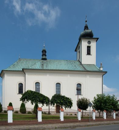 Zdjęcie nr 21: Kościół murowany, tynkowany, na kamiennym podmurowaniu, orientowany, jednonawowy. Prezbiterium prostokątne, krótkie, zamknięte trójbocznie, przy południowej ścianie prostokątna zakrystia z piętrem, z prostokątnym aneksem od zachodu. Nawa główna szersza od prezbiterium, na rzucie prostokąta, od zachodu przylega prostokątny przedsionek z wieżą na planie kwadratu. Wnętrze podzielone na cztery prostokątne przęsła i absydę, wydzielone arkadami, z gurtami spływającymi na zwielokrotnione pilastry. Każde przęsło nakryte sklepieniem o zaokrąglonych narożach. Łuk tęczowy półkolisty. W zachodniej części parapet chóru muzycznego wsparty na dwóch filarach i dwóch półfilarach, tworzących trzy arkady. Fasada trójosiowa, z wbudowaną wieżą, ujętą spłaszczonymi spływami zakończonymi cokołami z figurami. Osie wydzielone lizenami, podtrzymującymi profilowany gzyms, pod nim malowany fryz z dekoracją roślinną. Na osi portal w formie stojącego prostokąta zamkniętego łukiem odcinkowym, ujęty opaską tynkową, zakończoną kluczem. Drzwi drewniane, dwuskrzydłowe. Nad portalem fragment profilowanego gzymsu z daszkiem okapowym. Powyżej, na osi okno w formie stojącego prostokąta zamkniętego łukiem odcinkowym, w profilowanej opasce tynkowej, ujętej malowaną dekoracją geometryczną. Wieża dwukondygnacyjna, flankowana w narożach lizenami podtrzymującymi profilowany gzyms wieńczący. Na ścianie frontowej dwa okna na każdej kondygnacji, w formie stojącego prostokąta zamkniętego łukiem odcinkowym, w profilowanej opasce tynkowej, otoczonej malowaną dekoracją geometryczną, zwieńczone tarczą zegarową. Na pozostałych trzech ścianach wieży po jednym analogicznym oknie w drugiej kondygnacji i tarczy zegarowej. Elewacje północna i południowa artykułowane lizenami podtrzymującymi profilowany gzyms, z dekoracyjnym fryzem poniżej. Okna w formie stojącego prostokąta zamkniętego łukiem odcinkowym, w profilowanej opasce tynkowej z kluczem, ujętej malowaną dekoracją geometryczną: cztery w północnej i trzy w południowej. W zakrystii jedno prostokątne okno od południa i dwa w każdej kondygnacji od wschodu. Nawa główna i prezbiterium nakryte osobnymi dachami dwuspadowymi. Na ich połączeniu wieżyczka na sygnaturkę z latarnią, zwieńczona hełmem baniastym z krzyżem. Wieża nakryta hełmem baniastym zakończonym kulą z krzyżem. Zakrystia przykryta dachem trójspadowym, a aneks czterospadowym. 