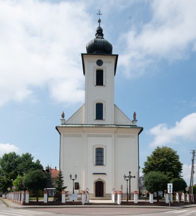 Zdjęcie nr 1: Kościół murowany, tynkowany, na kamiennym podmurowaniu, orientowany, jednonawowy. Prezbiterium prostokątne, krótkie, zamknięte trójbocznie, przy południowej ścianie prostokątna zakrystia z piętrem, z prostokątnym aneksem od zachodu. Nawa główna szersza od prezbiterium, na rzucie prostokąta, od zachodu przylega prostokątny przedsionek z wieżą na planie kwadratu. Wnętrze podzielone na cztery prostokątne przęsła i absydę, wydzielone arkadami, z gurtami spływającymi na zwielokrotnione pilastry. Każde przęsło nakryte sklepieniem o zaokrąglonych narożach. Łuk tęczowy półkolisty. W zachodniej części parapet chóru muzycznego wsparty na dwóch filarach i dwóch półfilarach, tworzących trzy arkady. Fasada trójosiowa, z wbudowaną wieżą, ujętą spłaszczonymi spływami zakończonymi cokołami z figurami. Osie wydzielone lizenami, podtrzymującymi profilowany gzyms, pod nim malowany fryz z dekoracją roślinną. Na osi portal w formie stojącego prostokąta zamkniętego łukiem odcinkowym, ujęty opaską tynkową, zakończoną kluczem. Drzwi drewniane, dwuskrzydłowe. Nad portalem fragment profilowanego gzymsu z daszkiem okapowym. Powyżej, na osi okno w formie stojącego prostokąta zamkniętego łukiem odcinkowym, w profilowanej opasce tynkowej, ujętej malowaną dekoracją geometryczną. Wieża dwukondygnacyjna, flankowana w narożach lizenami podtrzymującymi profilowany gzyms wieńczący. Na ścianie frontowej dwa okna na każdej kondygnacji, w formie stojącego prostokąta zamkniętego łukiem odcinkowym, w profilowanej opasce tynkowej, otoczonej malowaną dekoracją geometryczną, zwieńczone tarczą zegarową. Na pozostałych trzech ścianach wieży po jednym analogicznym oknie w drugiej kondygnacji i tarczy zegarowej. Elewacje północna i południowa artykułowane lizenami podtrzymującymi profilowany gzyms, z dekoracyjnym fryzem poniżej. Okna w formie stojącego prostokąta zamkniętego łukiem odcinkowym, w profilowanej opasce tynkowej z kluczem, ujętej malowaną dekoracją geometryczną: cztery w północnej i trzy w południowej. W zakrystii jedno prostokątne okno od południa i dwa w każdej kondygnacji od wschodu. Nawa główna i prezbiterium nakryte osobnymi dachami dwuspadowymi. Na ich połączeniu wieżyczka na sygnaturkę z latarnią, zwieńczona hełmem baniastym z krzyżem. Wieża nakryta hełmem baniastym zakończonym kulą z krzyżem. Zakrystia przykryta dachem trójspadowym, a aneks czterospadowym. 