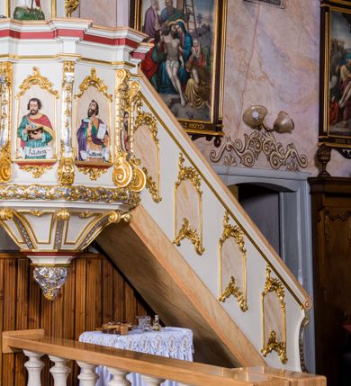 Zdjęcie nr 4: Ambona przyścienna, wisząca, z wejściem schodami od strony prezbiterium. Kosz jest na planie sześcioboku ze ściankami wydzielonymi pilastrami o trzonach w formie spływów wolutowych, zdobionych motywem  kampanuli i cekinów. Pola ścianek wypełniają płyciny w kształcie stojącego prostokąta dołem zamkniętego gzymsem,  górą łukiem wklęsło-wypukłym w formie ornamentu rocaille’owego. Umieszczono w nich płaskorzeźbione wizerunki czterech ewangelistów ukazanych w półpostaci. W dłoniach trzymają księgi lub zwoje oraz narzędzia pisarskie. U dołu każdej postaci znajduje się banderola z napisami (od lewej: „Św(ięty) Jan, Św(ięty) Łukasz, Św(ięty) Marek, Św(ięty) Mateusz”). Podstawę kosza obiega półwałek, natomiast podwieszenie, ujęte sześcioma spływami wolutowymi, zakończone jest dekoracyjną szyszką w kształcie osłoniętej liśćmi kiści winnej latorośli. Parapet ma formę pełnego belkowania wyłamującego się ponad wspierającymi go pilastrami.  Prowadzące na ambonę schody są jednobiegowe, z balustradą w formie ścianki zdobionej 4 płycinami w kształcie stojących prostokątów zamkniętych górą i dołem ornamentyką rocaille’ową, ujętej na końcu spływem wolutowym. Flankowany parą ażurowych, ornamentowych uszaków zaplecek wypełnia płycina w kształcie stojącego prostokąta zamkniętego łukiem wklęsło-wypukłym z płaskorzeźbionym, całopostaciowym przedstawieniem Chrystusa Dobrego Pasterza. Baldachim jest na rzucie ośmioboku, jego podstawę obiega wyłamujące się na narożach belkowanie, którego dolną krawędź zdobi motyw lambrekinu, natomiast górną kartusze rocaille’owe z kratką regencyjną. W podniebiu polichromowany motyw pokrytego obłokami nieba z płaskorzeźbioną gołębicą Ducha Świętego pośrodku. Naroża baldachimu ujmują spływy wolutowe pokryte ornamentem cekinowym. Zwieńczenie ma formę kopuły, na której osadzona jest figura dmącego w złoconą trąbę archanioła Michała stojącego na niebieskiej, ugwieżdżonej kuli, dołem ujętej wieńcem srebrzonymi obłoków. 
Struktura ambony utrzymana jest w kolorze białym, pola płycin są marmoryzowane w odcieniach kremowym i pomarańczowym, detal architektoniczny i ornamentyka są złocone i srebrzone oraz w kolorze bordowym.
