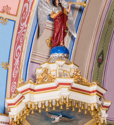 Zdjęcie nr 5: Ambona przyścienna, wisząca, z wejściem schodami od strony prezbiterium. Kosz jest na planie sześcioboku ze ściankami wydzielonymi pilastrami o trzonach w formie spływów wolutowych, zdobionych motywem  kampanuli i cekinów. Pola ścianek wypełniają płyciny w kształcie stojącego prostokąta dołem zamkniętego gzymsem,  górą łukiem wklęsło-wypukłym w formie ornamentu rocaille’owego. Umieszczono w nich płaskorzeźbione wizerunki czterech ewangelistów ukazanych w półpostaci. W dłoniach trzymają księgi lub zwoje oraz narzędzia pisarskie. U dołu każdej postaci znajduje się banderola z napisami (od lewej: „Św(ięty) Jan, Św(ięty) Łukasz, Św(ięty) Marek, Św(ięty) Mateusz”). Podstawę kosza obiega półwałek, natomiast podwieszenie, ujęte sześcioma spływami wolutowymi, zakończone jest dekoracyjną szyszką w kształcie osłoniętej liśćmi kiści winnej latorośli. Parapet ma formę pełnego belkowania wyłamującego się ponad wspierającymi go pilastrami.  Prowadzące na ambonę schody są jednobiegowe, z balustradą w formie ścianki zdobionej 4 płycinami w kształcie stojących prostokątów zamkniętych górą i dołem ornamentyką rocaille’ową, ujętej na końcu spływem wolutowym. Flankowany parą ażurowych, ornamentowych uszaków zaplecek wypełnia płycina w kształcie stojącego prostokąta zamkniętego łukiem wklęsło-wypukłym z płaskorzeźbionym, całopostaciowym przedstawieniem Chrystusa Dobrego Pasterza. Baldachim jest na rzucie ośmioboku, jego podstawę obiega wyłamujące się na narożach belkowanie, którego dolną krawędź zdobi motyw lambrekinu, natomiast górną kartusze rocaille’owe z kratką regencyjną. W podniebiu polichromowany motyw pokrytego obłokami nieba z płaskorzeźbioną gołębicą Ducha Świętego pośrodku. Naroża baldachimu ujmują spływy wolutowe pokryte ornamentem cekinowym. Zwieńczenie ma formę kopuły, na której osadzona jest figura dmącego w złoconą trąbę archanioła Michała stojącego na niebieskiej, ugwieżdżonej kuli, dołem ujętej wieńcem srebrzonymi obłoków. 
Struktura ambony utrzymana jest w kolorze białym, pola płycin są marmoryzowane w odcieniach kremowym i pomarańczowym, detal architektoniczny i ornamentyka są złocone i srebrzone oraz w kolorze bordowym.
