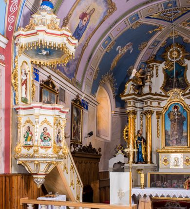 Zdjęcie nr 2: Ambona przyścienna, wisząca, z wejściem schodami od strony prezbiterium. Kosz jest na planie sześcioboku ze ściankami wydzielonymi pilastrami o trzonach w formie spływów wolutowych, zdobionych motywem  kampanuli i cekinów. Pola ścianek wypełniają płyciny w kształcie stojącego prostokąta dołem zamkniętego gzymsem,  górą łukiem wklęsło-wypukłym w formie ornamentu rocaille’owego. Umieszczono w nich płaskorzeźbione wizerunki czterech ewangelistów ukazanych w półpostaci. W dłoniach trzymają księgi lub zwoje oraz narzędzia pisarskie. U dołu każdej postaci znajduje się banderola z napisami (od lewej: „Św(ięty) Jan, Św(ięty) Łukasz, Św(ięty) Marek, Św(ięty) Mateusz”). Podstawę kosza obiega półwałek, natomiast podwieszenie, ujęte sześcioma spływami wolutowymi, zakończone jest dekoracyjną szyszką w kształcie osłoniętej liśćmi kiści winnej latorośli. Parapet ma formę pełnego belkowania wyłamującego się ponad wspierającymi go pilastrami.  Prowadzące na ambonę schody są jednobiegowe, z balustradą w formie ścianki zdobionej 4 płycinami w kształcie stojących prostokątów zamkniętych górą i dołem ornamentyką rocaille’ową, ujętej na końcu spływem wolutowym. Flankowany parą ażurowych, ornamentowych uszaków zaplecek wypełnia płycina w kształcie stojącego prostokąta zamkniętego łukiem wklęsło-wypukłym z płaskorzeźbionym, całopostaciowym przedstawieniem Chrystusa Dobrego Pasterza. Baldachim jest na rzucie ośmioboku, jego podstawę obiega wyłamujące się na narożach belkowanie, którego dolną krawędź zdobi motyw lambrekinu, natomiast górną kartusze rocaille’owe z kratką regencyjną. W podniebiu polichromowany motyw pokrytego obłokami nieba z płaskorzeźbioną gołębicą Ducha Świętego pośrodku. Naroża baldachimu ujmują spływy wolutowe pokryte ornamentem cekinowym. Zwieńczenie ma formę kopuły, na której osadzona jest figura dmącego w złoconą trąbę archanioła Michała stojącego na niebieskiej, ugwieżdżonej kuli, dołem ujętej wieńcem srebrzonymi obłoków. 
Struktura ambony utrzymana jest w kolorze białym, pola płycin są marmoryzowane w odcieniach kremowym i pomarańczowym, detal architektoniczny i ornamentyka są złocone i srebrzone oraz w kolorze bordowym.
