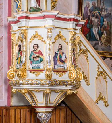 Zdjęcie nr 3: Ambona przyścienna, wisząca, z wejściem schodami od strony prezbiterium. Kosz jest na planie sześcioboku ze ściankami wydzielonymi pilastrami o trzonach w formie spływów wolutowych, zdobionych motywem  kampanuli i cekinów. Pola ścianek wypełniają płyciny w kształcie stojącego prostokąta dołem zamkniętego gzymsem,  górą łukiem wklęsło-wypukłym w formie ornamentu rocaille’owego. Umieszczono w nich płaskorzeźbione wizerunki czterech ewangelistów ukazanych w półpostaci. W dłoniach trzymają księgi lub zwoje oraz narzędzia pisarskie. U dołu każdej postaci znajduje się banderola z napisami (od lewej: „Św(ięty) Jan, Św(ięty) Łukasz, Św(ięty) Marek, Św(ięty) Mateusz”). Podstawę kosza obiega półwałek, natomiast podwieszenie, ujęte sześcioma spływami wolutowymi, zakończone jest dekoracyjną szyszką w kształcie osłoniętej liśćmi kiści winnej latorośli. Parapet ma formę pełnego belkowania wyłamującego się ponad wspierającymi go pilastrami.  Prowadzące na ambonę schody są jednobiegowe, z balustradą w formie ścianki zdobionej 4 płycinami w kształcie stojących prostokątów zamkniętych górą i dołem ornamentyką rocaille’ową, ujętej na końcu spływem wolutowym. Flankowany parą ażurowych, ornamentowych uszaków zaplecek wypełnia płycina w kształcie stojącego prostokąta zamkniętego łukiem wklęsło-wypukłym z płaskorzeźbionym, całopostaciowym przedstawieniem Chrystusa Dobrego Pasterza. Baldachim jest na rzucie ośmioboku, jego podstawę obiega wyłamujące się na narożach belkowanie, którego dolną krawędź zdobi motyw lambrekinu, natomiast górną kartusze rocaille’owe z kratką regencyjną. W podniebiu polichromowany motyw pokrytego obłokami nieba z płaskorzeźbioną gołębicą Ducha Świętego pośrodku. Naroża baldachimu ujmują spływy wolutowe pokryte ornamentem cekinowym. Zwieńczenie ma formę kopuły, na której osadzona jest figura dmącego w złoconą trąbę archanioła Michała stojącego na niebieskiej, ugwieżdżonej kuli, dołem ujętej wieńcem srebrzonymi obłoków. 
Struktura ambony utrzymana jest w kolorze białym, pola płycin są marmoryzowane w odcieniach kremowym i pomarańczowym, detal architektoniczny i ornamentyka są złocone i srebrzone oraz w kolorze bordowym.
