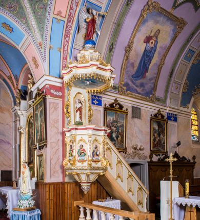 Zdjęcie nr 1: Ambona przyścienna, wisząca, z wejściem schodami od strony prezbiterium. Kosz jest na planie sześcioboku ze ściankami wydzielonymi pilastrami o trzonach w formie spływów wolutowych, zdobionych motywem  kampanuli i cekinów. Pola ścianek wypełniają płyciny w kształcie stojącego prostokąta dołem zamkniętego gzymsem,  górą łukiem wklęsło-wypukłym w formie ornamentu rocaille’owego. Umieszczono w nich płaskorzeźbione wizerunki czterech ewangelistów ukazanych w półpostaci. W dłoniach trzymają księgi lub zwoje oraz narzędzia pisarskie. U dołu każdej postaci znajduje się banderola z napisami (od lewej: „Św(ięty) Jan, Św(ięty) Łukasz, Św(ięty) Marek, Św(ięty) Mateusz”). Podstawę kosza obiega półwałek, natomiast podwieszenie, ujęte sześcioma spływami wolutowymi, zakończone jest dekoracyjną szyszką w kształcie osłoniętej liśćmi kiści winnej latorośli. Parapet ma formę pełnego belkowania wyłamującego się ponad wspierającymi go pilastrami.  Prowadzące na ambonę schody są jednobiegowe, z balustradą w formie ścianki zdobionej 4 płycinami w kształcie stojących prostokątów zamkniętych górą i dołem ornamentyką rocaille’ową, ujętej na końcu spływem wolutowym. Flankowany parą ażurowych, ornamentowych uszaków zaplecek wypełnia płycina w kształcie stojącego prostokąta zamkniętego łukiem wklęsło-wypukłym z płaskorzeźbionym, całopostaciowym przedstawieniem Chrystusa Dobrego Pasterza. Baldachim jest na rzucie ośmioboku, jego podstawę obiega wyłamujące się na narożach belkowanie, którego dolną krawędź zdobi motyw lambrekinu, natomiast górną kartusze rocaille’owe z kratką regencyjną. W podniebiu polichromowany motyw pokrytego obłokami nieba z płaskorzeźbioną gołębicą Ducha Świętego pośrodku. Naroża baldachimu ujmują spływy wolutowe pokryte ornamentem cekinowym. Zwieńczenie ma formę kopuły, na której osadzona jest figura dmącego w złoconą trąbę archanioła Michała stojącego na niebieskiej, ugwieżdżonej kuli, dołem ujętej wieńcem srebrzonymi obłoków. 
Struktura ambony utrzymana jest w kolorze białym, pola płycin są marmoryzowane w odcieniach kremowym i pomarańczowym, detal architektoniczny i ornamentyka są złocone i srebrzone oraz w kolorze bordowym.
