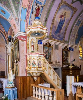 Zdjęcie nr 1: Ambona przyścienna, wisząca, z wejściem schodami od strony prezbiterium. Kosz jest na planie sześcioboku ze ściankami wydzielonymi pilastrami o trzonach w formie spływów wolutowych, zdobionych motywem  kampanuli i cekinów. Pola ścianek wypełniają płyciny w kształcie stojącego prostokąta dołem zamkniętego gzymsem,  górą łukiem wklęsło-wypukłym w formie ornamentu rocaille’owego. Umieszczono w nich płaskorzeźbione wizerunki czterech ewangelistów ukazanych w półpostaci. W dłoniach trzymają księgi lub zwoje oraz narzędzia pisarskie. U dołu każdej postaci znajduje się banderola z napisami (od lewej: „Św(ięty) Jan, Św(ięty) Łukasz, Św(ięty) Marek, Św(ięty) Mateusz”). Podstawę kosza obiega półwałek, natomiast podwieszenie, ujęte sześcioma spływami wolutowymi, zakończone jest dekoracyjną szyszką w kształcie osłoniętej liśćmi kiści winnej latorośli. Parapet ma formę pełnego belkowania wyłamującego się ponad wspierającymi go pilastrami.  Prowadzące na ambonę schody są jednobiegowe, z balustradą w formie ścianki zdobionej 4 płycinami w kształcie stojących prostokątów zamkniętych górą i dołem ornamentyką rocaille’ową, ujętej na końcu spływem wolutowym. Flankowany parą ażurowych, ornamentowych uszaków zaplecek wypełnia płycina w kształcie stojącego prostokąta zamkniętego łukiem wklęsło-wypukłym z płaskorzeźbionym, całopostaciowym przedstawieniem Chrystusa Dobrego Pasterza. Baldachim jest na rzucie ośmioboku, jego podstawę obiega wyłamujące się na narożach belkowanie, którego dolną krawędź zdobi motyw lambrekinu, natomiast górną kartusze rocaille’owe z kratką regencyjną. W podniebiu polichromowany motyw pokrytego obłokami nieba z płaskorzeźbioną gołębicą Ducha Świętego pośrodku. Naroża baldachimu ujmują spływy wolutowe pokryte ornamentem cekinowym. Zwieńczenie ma formę kopuły, na której osadzona jest figura dmącego w złoconą trąbę archanioła Michała stojącego na niebieskiej, ugwieżdżonej kuli, dołem ujętej wieńcem srebrzonymi obłoków. 
Struktura ambony utrzymana jest w kolorze białym, pola płycin są marmoryzowane w odcieniach kremowym i pomarańczowym, detal architektoniczny i ornamentyka są złocone i srebrzone oraz w kolorze bordowym.
