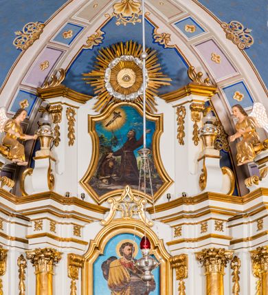 Zdjęcie nr 4: Ołtarz architektoniczny, jednokondygnacyjny, jednoosiowy, ze zwieńczeniem. Ołtarz murowany, prostopadłościenny z antepedium w formie prostokątnego obrazu ze sceną Ostatniej Wieczerzy według fresku Leonarda da Vinci. Na mensie tabernakulum w formie prostopadłościennej skrzyni osadzonej na niewielkim cokole i zwieńczonej gzymsem zdobionym wicią winnej latorośli. Metalowe drzwiczki w kształcie stojącego prostokąta z płyciną z motywem kielicha eucharystycznego, po bokach zdobione wicią winnej latorośli, flankowane parą prostokątnych płycin z motywami płaskorzeźbionych, uskrzydlonych główek anielskich. Na tabernakulum po bokach para figur klęczących aniołów w długich, złoconych sukniach. Retabulum osadzone na wysokim zryzalitowanym ukośnie po bokach cokole. Część środkowa ujęta dwoma parami kolumn ustawionych na tle dwóch par filarów, z czego skrajne bardziej wysunięte. Podpory wspierają wyłamujące się ponad nimi przerwane belkowanie. Zwieńczenie ujęte parą ukośnie ustawionych spływów wolutowych, zamknięte łukiem odcinkowym, flankowane przerwanym wolutowym przyczółkiem. Po bokach retabulum para bramek o przejściach zamkniętych łukami kotarowymi, zwieńczonych fragmentami gzymsów w postaci łuków wklęsło-wypukłych. Trzony filarów zdobione kampanulami, fryz rozetami. Retabulum ujmuje para ażurowych uszaków. Pośrodku przerwanego belkowania monogram maryjny. Pola spływów wolutowych zwieńczenia zdobią motywy kampanuli, szczyt w formie promienistej glorii z okiem Opatrzności pośrodku ujętym kręgiem obłoków. Na przerwanym przyczółku para figur siedzących aniołów w długich, złoconych sukniach. Pole części środkowej w kształcie stojącego prostokąta zamkniętego półkolistym łukiem nadwieszonym z uskokami, ujęte profilowaną, złoconą ramą. W środku obraz Matki Boskiej Pocieszenia, na zasuwie wizerunek św. Jakuba Starszego. Po bokach, między kolumnami para figur, po prawej stronie św. Paweł, po lewej św. Piotr. W zwieńczeniu obraz w kształcie trapezu o wklęsłych ramionach i podstawie, ściętych narożach dolnych, zamknięty łukiem wklęsło-wypukłym z przedstawieniem stygmatyzacji św. Franciszka. Struktura polichromowana w kolorze białym, częściowo marmoryzowana w odcieniach beżów, detal architektoniczny i ornamentyka złocone, marmoryzowane i srebrzone.     