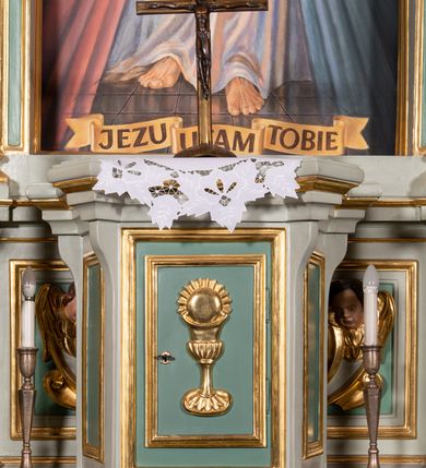 Zdjęcie nr 18: Ołtarz przyścienny, prostopadłościenny, z antepedium, na którym umieszczone są rzeźbione litery IHS; mensa drewniana z kamiennym portatylem. Na mensie znajduje się tabernakulum o kształcie graniastosłupa, z drzwiczkami ozdobionymi rzeźbionym kielichem z hostią. Nastawa ołtarzowa jest architektoniczna, jednoosiowa, jednokondygnacyjna ze zwieńczeniem. Osadzona na podwójnym postumencie, flankowana pilastrami o kompozytowych kapitelach i kanelowanymi kolumnami o kompozytowych kapitelach, które podtrzymują belkowanie. Ponad belkowaniem znajduje się przełamany przyczółek w formie łuku półkolistego, bogato profilowany, ozdobiony ulistnionymi konsolkami i rozetami oraz girlandami z owoców. W partii fryzu kartusz o fantazyjnym wykroju, ujęty wolutami i listkami akantu. Na belkowaniu siedzą po bokach nagie putta, odziane w opaski na biodrach, a pośrodku znajduje się krzyż osadzony na postumencie z liśćmi. Po obu stronach ołtarza na konsolach o krawędziach ozdobionych ornamentem małżowinowym stoją figury trudnych do identyfikacji świętych, być może apostołów. W centralnej części ołtarza wnęka prostokątna, zamknięta łukiem półkolistym. W niej obraz z przedstawieniem „Jezu ufam Tobie”. 