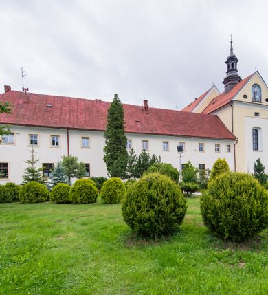 Zdjęcie nr 27: Kompleks klasztorny jest na rzucie czworoboku, z wirydarzem pośrodku i kościołem przylegającym od wschodu. Kościół, zwrócony w kierunku południowo-wschodnim, jest budowlą murowaną z cegły i kamienia, na rzucie podłużnym. Korpus jest jednonawowy i dwuprzęsłowy. Do dwuprzęsłowego prezbiterium na rzucie prostokąta od wschodu przylega zakrystia, do korpusu nawowego kaplica na rzucie ośmioboku z niewysoką przyległością, natomiast do fasady kruchta. Nawa i prezbiterium przekryte są sklepieniami kolebkowymi z lunetami wspartymi na masywnych półfilarach. Przestrzeń prezbiterium od korpusu nawowego oddziela półkolista arkada łuku tęczowego. Artykulację ścian na zewnątrz kościoła stanowią ujmujące okna duże, prostokątne płyciny. Korpus i prezbiterium obiega gzyms koronujący. Szczyt kościoła trójkątny, kruchty w formie nadwieszonego łuku z wycinka elipsy z wklęsłymi uskokami. Do wnętrza prowadzą dwa otwory drzwiowe po wschodniej i północnej stronie kruchty z parą prostokątnych, dwuskrzydłowych drzwi. W szczycie fasady płytka wnęka w kształcie stojącego prostokąta zamkniętego półkoliście z wizerunkiem Niepokalanego Poczęcia Najświętszej Marii Panny, ujęta trzema okrągłymi oknami. W szczycie kruchty analogiczna wnęka z figurą św. Floriana. Pośrodku fasady duże okno w kształcie stojącego prostokąta zamkniętego łukiem odcinkowym. Analogiczne okna w przęsłach korpusu i prezbiterium oraz na jego zamknięciu. Szczyt prezbiterium artykułuje płytka wnęka w kształcie stojącego prostokąta zamkniętego półkoliście z wizerunkiem św. Franciszka adorującego Jezusa na krzyżu flankowana parą okrągłych okien. Korpus i prezbiterium przekryte dachami dwuspadowymi, nad prezbiterium wieżyczka na sygnaturkę. Ściany kaplicy bocznej w formie półkolistych arkad, wypełnionych dużymi oknami z dekoracją maswerkową w górnej partii i przeszklonym otworem drzwiowym, zwieńczonych trójkątnymi szczytami. Ujmują je pary pilastrów, szczyty artykułuje fryz arkadkowy. Kaplicę wieńczy hełm pinaklowy z latarnią. Ściany sąsiedniej przyległości, nakrytej dachem pulpitowym i zakrystii z dachem trójspadowym, artykułuje fryz arkadkowy z lizenami. Tynki pomalowane są w odcieniach żółci. 