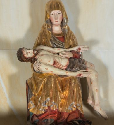 Zdjęcie nr 1: Całopostaciowa, ścięta z tyłu figura przedstawiająca Pietę. Ukazana frontalnie Maria siedzi na prostopadłościennym stołku. Nieznacznie przechyloną w prawo i lekko pochyloną głowę charakteryzują pociągła twarz o owalnym obrysie i subtelnych rysach twarzy, cienkie linie łagodnych łuków brwi, niewielkie oczy o migdałowatym kształcie, długi, prosty nos, drobne usta o lekko opadających kącikach, policzki i podbródek podkreślone rumieńcem. Maria ma na sobie czerwoną suknię z szerokim, złoconym obszyciem dekoltu oraz obszerny, złocony płaszcz o srebrzonym podszyciu, osłaniający głowę, ręce i nogi, o prawej pole podwiniętej pod lewą rękę. Fałdowania przybierają formę drobnych rurek opadających kaskadowo po bokach twarzy oraz w dolnej partii nóg, na wysokości łydek załamujących się w ostre formy w kształcie litery „V”. Maria oburącz podtrzymuje leżące na jej kolanach martwe ciało Jezusa, o zwróconej w lewo sylwetce. Trójkątną twarz charakteryzują wyraziste rysy z niskim czołem, cienką, prostą linią brwi, zamkniętymi oczami, długim, prostym wydatnym nosem, wydatnymi kośćmi policzkowymi, krótkim zarostem. Długie, lekko falowane, brązowe włosy opadają na plecy. Sztywną sylwetkę cechuje także sumarycznie modelowana muskulatura wydatna zwłaszcza w partii brzucha. Długie wyprostowane ręce ułożone są wzdłuż sylwetki, lewa wsparta na lewej ręce Marii; dłonie duże, o wyprostowanych palcach. Długie, zgięte w kolanach nogi o dużych stopach opadają prostopadle do podłoża. Biodra Jezusa przesłania krótkie, ciemnoszare perizonium. Całe ciało o bladej karnacji pokrywają drobne strużki krwi składające się z trzech wydłużonych kropli, na stopach, dłoniach i w prawym boku wydatne, mocno krwawiące rany, czoło wzdłuż linii włosów mocno zakrwawione.  