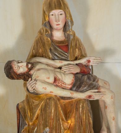 Zdjęcie nr 3: Całopostaciowa, ścięta z tyłu figura przedstawiająca Pietę. Ukazana frontalnie Maria siedzi na prostopadłościennym stołku. Nieznacznie przechyloną w prawo i lekko pochyloną głowę charakteryzują pociągła twarz o owalnym obrysie i subtelnych rysach twarzy, cienkie linie łagodnych łuków brwi, niewielkie oczy o migdałowatym kształcie, długi, prosty nos, drobne usta o lekko opadających kącikach, policzki i podbródek podkreślone rumieńcem. Maria ma na sobie czerwoną suknię z szerokim, złoconym obszyciem dekoltu oraz obszerny, złocony płaszcz o srebrzonym podszyciu, osłaniający głowę, ręce i nogi, o prawej pole podwiniętej pod lewą rękę. Fałdowania przybierają formę drobnych rurek opadających kaskadowo po bokach twarzy oraz w dolnej partii nóg, na wysokości łydek załamujących się w ostre formy w kształcie litery „V”. Maria oburącz podtrzymuje leżące na jej kolanach martwe ciało Jezusa, o zwróconej w lewo sylwetce. Trójkątną twarz charakteryzują wyraziste rysy z niskim czołem, cienką, prostą linią brwi, zamkniętymi oczami, długim, prostym wydatnym nosem, wydatnymi kośćmi policzkowymi, krótkim zarostem. Długie, lekko falowane, brązowe włosy opadają na plecy. Sztywną sylwetkę cechuje także sumarycznie modelowana muskulatura wydatna zwłaszcza w partii brzucha. Długie wyprostowane ręce ułożone są wzdłuż sylwetki, lewa wsparta na lewej ręce Marii; dłonie duże, o wyprostowanych palcach. Długie, zgięte w kolanach nogi o dużych stopach opadają prostopadle do podłoża. Biodra Jezusa przesłania krótkie, ciemnoszare perizonium. Całe ciało o bladej karnacji pokrywają drobne strużki krwi składające się z trzech wydłużonych kropli, na stopach, dłoniach i w prawym boku wydatne, mocno krwawiące rany, czoło wzdłuż linii włosów mocno zakrwawione.  