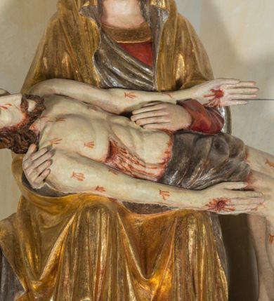 Zdjęcie nr 4: Całopostaciowa, ścięta z tyłu figura przedstawiająca Pietę. Ukazana frontalnie Maria siedzi na prostopadłościennym stołku. Nieznacznie przechyloną w prawo i lekko pochyloną głowę charakteryzują pociągła twarz o owalnym obrysie i subtelnych rysach twarzy, cienkie linie łagodnych łuków brwi, niewielkie oczy o migdałowatym kształcie, długi, prosty nos, drobne usta o lekko opadających kącikach, policzki i podbródek podkreślone rumieńcem. Maria ma na sobie czerwoną suknię z szerokim, złoconym obszyciem dekoltu oraz obszerny, złocony płaszcz o srebrzonym podszyciu, osłaniający głowę, ręce i nogi, o prawej pole podwiniętej pod lewą rękę. Fałdowania przybierają formę drobnych rurek opadających kaskadowo po bokach twarzy oraz w dolnej partii nóg, na wysokości łydek załamujących się w ostre formy w kształcie litery „V”. Maria oburącz podtrzymuje leżące na jej kolanach martwe ciało Jezusa, o zwróconej w lewo sylwetce. Trójkątną twarz charakteryzują wyraziste rysy z niskim czołem, cienką, prostą linią brwi, zamkniętymi oczami, długim, prostym wydatnym nosem, wydatnymi kośćmi policzkowymi, krótkim zarostem. Długie, lekko falowane, brązowe włosy opadają na plecy. Sztywną sylwetkę cechuje także sumarycznie modelowana muskulatura wydatna zwłaszcza w partii brzucha. Długie wyprostowane ręce ułożone są wzdłuż sylwetki, lewa wsparta na lewej ręce Marii; dłonie duże, o wyprostowanych palcach. Długie, zgięte w kolanach nogi o dużych stopach opadają prostopadle do podłoża. Biodra Jezusa przesłania krótkie, ciemnoszare perizonium. Całe ciało o bladej karnacji pokrywają drobne strużki krwi składające się z trzech wydłużonych kropli, na stopach, dłoniach i w prawym boku wydatne, mocno krwawiące rany, czoło wzdłuż linii włosów mocno zakrwawione.  