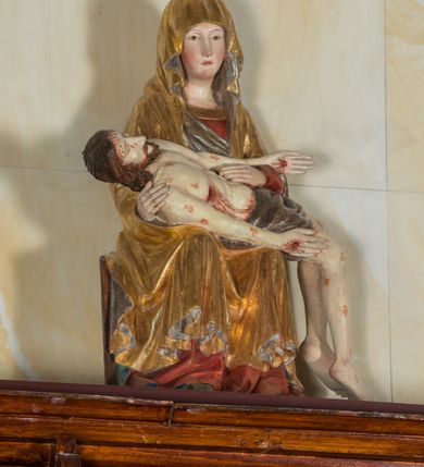 Zdjęcie nr 2: Całopostaciowa, ścięta z tyłu figura przedstawiająca Pietę. Ukazana frontalnie Maria siedzi na prostopadłościennym stołku. Nieznacznie przechyloną w prawo i lekko pochyloną głowę charakteryzują pociągła twarz o owalnym obrysie i subtelnych rysach twarzy, cienkie linie łagodnych łuków brwi, niewielkie oczy o migdałowatym kształcie, długi, prosty nos, drobne usta o lekko opadających kącikach, policzki i podbródek podkreślone rumieńcem. Maria ma na sobie czerwoną suknię z szerokim, złoconym obszyciem dekoltu oraz obszerny, złocony płaszcz o srebrzonym podszyciu, osłaniający głowę, ręce i nogi, o prawej pole podwiniętej pod lewą rękę. Fałdowania przybierają formę drobnych rurek opadających kaskadowo po bokach twarzy oraz w dolnej partii nóg, na wysokości łydek załamujących się w ostre formy w kształcie litery „V”. Maria oburącz podtrzymuje leżące na jej kolanach martwe ciało Jezusa, o zwróconej w lewo sylwetce. Trójkątną twarz charakteryzują wyraziste rysy z niskim czołem, cienką, prostą linią brwi, zamkniętymi oczami, długim, prostym wydatnym nosem, wydatnymi kośćmi policzkowymi, krótkim zarostem. Długie, lekko falowane, brązowe włosy opadają na plecy. Sztywną sylwetkę cechuje także sumarycznie modelowana muskulatura wydatna zwłaszcza w partii brzucha. Długie wyprostowane ręce ułożone są wzdłuż sylwetki, lewa wsparta na lewej ręce Marii; dłonie duże, o wyprostowanych palcach. Długie, zgięte w kolanach nogi o dużych stopach opadają prostopadle do podłoża. Biodra Jezusa przesłania krótkie, ciemnoszare perizonium. Całe ciało o bladej karnacji pokrywają drobne strużki krwi składające się z trzech wydłużonych kropli, na stopach, dłoniach i w prawym boku wydatne, mocno krwawiące rany, czoło wzdłuż linii włosów mocno zakrwawione.  