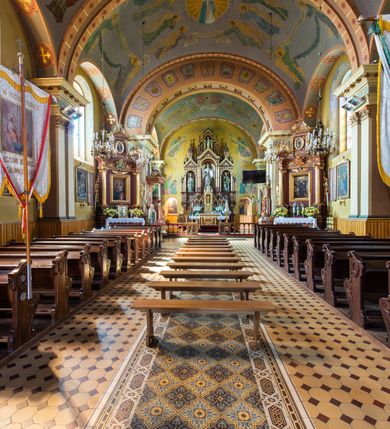 Zdjęcie nr 9: Kościół, zwrócony w kierunku południowo-wschodnim, jest budowlą murowaną z cegły na zaprawie wapiennej, na rzucie podłużnym. Powierzchnia budynku wynosi około 550 m2. Kościół charakteryzuje zwarta, prosta bryła. Korpus jest jednonawowy i trójprzęsłowy. Szerokie, dwuprzęsłowe, prezbiterium jest zamknięte półkoliście. Na osi kościoła, w przedłużeniu północnego przęsła przylega jednokondygnacyjna wieża, ujęta po bokach piętrowymi lokalnościami. Nawa przekryta jest sklepieniem kolebkowym z gurtami, wspartymi na osadzonych na wysokich cokołach parach przyściennych, czworobocznych filarów z przylegającymi do nich pilastrami o gładkich trzonach. Przestrzeń prezbiterium od korpusu nawowego oddziela półkoliście zamknięta arkada łuku tęczowego i drewniana balustrada. Zamknięte prostą ścianą prezbiterium zajmuje przestrzeń jednego przęsła. Dalej znajduje się zakrystia. Artykulację ścian na zewnątrz kościoła stanowi niewysoki obiegający bryłę cokół oraz niepełne belkowanie wieńczące, składające się z fryzu i gzymsu. Fasadę artykułuje płycina podzielona parą wspartych na cokołach pilastrów. W polu środkowym znajduje się główny otwór wejściowy. Dwuskrzydłowe, drewniane drzwi z naświetlem ujęte są prostym, profilowanym portalem. Tuż nad nadprożem znajduje się krótki gzyms. Wyżej duże, prostokątne, zamknięte półkoliście okno. Fasadę wieńczy trójkątny przyczółek. Ściany korpusu nawowego i prezbiterium przeprute są prostokątnymi, zamkniętymi półkoliście oknami. Dodatkowo w ścianie zachodniej, na wysokości przyległości wieży, w dolnej partii muru znajduje się niewielkie, półkoliście zamknięte okno a w drugim przęśle nawy prostokątny otwór drzwiowy; podobny jest umiejscowiony w drugim przęśle prezbiterium po stronie zachodniej. Kondygnację czworobocznej wieży w dolnej partii obiega parapet, wyżej w każdej ze ścian znajduje się po jednym prostokątnym, zamkniętym półkoliście oknie. Górną partię wieży obiega gzyms wieńczący wyłamujący się półkoliście pośrodku każdej ze ścian. Wieża nakryta jest hełmem cebulastym z latarnią. Korpus nawowy i prezbiterium przekrywa dwuspadowy dach wykonany z blachy, z wieżyczką na sygnaturkę na zakończeniu prezbiterium. Kolorystyka tynków kościoła utrzymana jest w odcieniach żółci i bieli, cokół jest brązowy.  Okna nawy, prezbiterium i fasady wypełniają witraże, wnętrze pokrywa polichromia z przedstawieniami figuralnymi w partii sklepień. Plac przykościelny, z wybrukowanymi alejkami i trawnikiem, otoczony jest żywopłotem z krzewów ozdobnych.