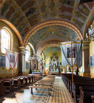 Zdjęcie nr 13: Kościół, zwrócony w kierunku południowo-wschodnim, jest budowlą murowaną z cegły na zaprawie wapiennej, na rzucie podłużnym. Powierzchnia budynku wynosi około 550 m2. Kościół charakteryzuje zwarta, prosta bryła. Korpus jest jednonawowy i trójprzęsłowy. Szerokie, dwuprzęsłowe, prezbiterium jest zamknięte półkoliście. Na osi kościoła, w przedłużeniu północnego przęsła przylega jednokondygnacyjna wieża, ujęta po bokach piętrowymi lokalnościami. Nawa przekryta jest sklepieniem kolebkowym z gurtami, wspartymi na osadzonych na wysokich cokołach parach przyściennych, czworobocznych filarów z przylegającymi do nich pilastrami o gładkich trzonach. Przestrzeń prezbiterium od korpusu nawowego oddziela półkoliście zamknięta arkada łuku tęczowego i drewniana balustrada. Zamknięte prostą ścianą prezbiterium zajmuje przestrzeń jednego przęsła. Dalej znajduje się zakrystia. Artykulację ścian na zewnątrz kościoła stanowi niewysoki obiegający bryłę cokół oraz niepełne belkowanie wieńczące, składające się z fryzu i gzymsu. Fasadę artykułuje płycina podzielona parą wspartych na cokołach pilastrów. W polu środkowym znajduje się główny otwór wejściowy. Dwuskrzydłowe, drewniane drzwi z naświetlem ujęte są prostym, profilowanym portalem. Tuż nad nadprożem znajduje się krótki gzyms. Wyżej duże, prostokątne, zamknięte półkoliście okno. Fasadę wieńczy trójkątny przyczółek. Ściany korpusu nawowego i prezbiterium przeprute są prostokątnymi, zamkniętymi półkoliście oknami. Dodatkowo w ścianie zachodniej, na wysokości przyległości wieży, w dolnej partii muru znajduje się niewielkie, półkoliście zamknięte okno a w drugim przęśle nawy prostokątny otwór drzwiowy; podobny jest umiejscowiony w drugim przęśle prezbiterium po stronie zachodniej. Kondygnację czworobocznej wieży w dolnej partii obiega parapet, wyżej w każdej ze ścian znajduje się po jednym prostokątnym, zamkniętym półkoliście oknie. Górną partię wieży obiega gzyms wieńczący wyłamujący się półkoliście pośrodku każdej ze ścian. Wieża nakryta jest hełmem cebulastym z latarnią. Korpus nawowy i prezbiterium przekrywa dwuspadowy dach wykonany z blachy, z wieżyczką na sygnaturkę na zakończeniu prezbiterium. Kolorystyka tynków kościoła utrzymana jest w odcieniach żółci i bieli, cokół jest brązowy.  Okna nawy, prezbiterium i fasady wypełniają witraże, wnętrze pokrywa polichromia z przedstawieniami figuralnymi w partii sklepień. Plac przykościelny, z wybrukowanymi alejkami i trawnikiem, otoczony jest żywopłotem z krzewów ozdobnych.