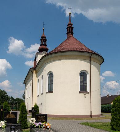 Zdjęcie nr 6: Kościół, zwrócony w kierunku południowo-wschodnim, jest budowlą murowaną z cegły na zaprawie wapiennej, na rzucie podłużnym. Powierzchnia budynku wynosi około 550 m2. Kościół charakteryzuje zwarta, prosta bryła. Korpus jest jednonawowy i trójprzęsłowy. Szerokie, dwuprzęsłowe, prezbiterium jest zamknięte półkoliście. Na osi kościoła, w przedłużeniu północnego przęsła przylega jednokondygnacyjna wieża, ujęta po bokach piętrowymi lokalnościami. Nawa przekryta jest sklepieniem kolebkowym z gurtami, wspartymi na osadzonych na wysokich cokołach parach przyściennych, czworobocznych filarów z przylegającymi do nich pilastrami o gładkich trzonach. Przestrzeń prezbiterium od korpusu nawowego oddziela półkoliście zamknięta arkada łuku tęczowego i drewniana balustrada. Zamknięte prostą ścianą prezbiterium zajmuje przestrzeń jednego przęsła. Dalej znajduje się zakrystia. Artykulację ścian na zewnątrz kościoła stanowi niewysoki obiegający bryłę cokół oraz niepełne belkowanie wieńczące, składające się z fryzu i gzymsu. Fasadę artykułuje płycina podzielona parą wspartych na cokołach pilastrów. W polu środkowym znajduje się główny otwór wejściowy. Dwuskrzydłowe, drewniane drzwi z naświetlem ujęte są prostym, profilowanym portalem. Tuż nad nadprożem znajduje się krótki gzyms. Wyżej duże, prostokątne, zamknięte półkoliście okno. Fasadę wieńczy trójkątny przyczółek. Ściany korpusu nawowego i prezbiterium przeprute są prostokątnymi, zamkniętymi półkoliście oknami. Dodatkowo w ścianie zachodniej, na wysokości przyległości wieży, w dolnej partii muru znajduje się niewielkie, półkoliście zamknięte okno a w drugim przęśle nawy prostokątny otwór drzwiowy; podobny jest umiejscowiony w drugim przęśle prezbiterium po stronie zachodniej. Kondygnację czworobocznej wieży w dolnej partii obiega parapet, wyżej w każdej ze ścian znajduje się po jednym prostokątnym, zamkniętym półkoliście oknie. Górną partię wieży obiega gzyms wieńczący wyłamujący się półkoliście pośrodku każdej ze ścian. Wieża nakryta jest hełmem cebulastym z latarnią. Korpus nawowy i prezbiterium przekrywa dwuspadowy dach wykonany z blachy, z wieżyczką na sygnaturkę na zakończeniu prezbiterium. Kolorystyka tynków kościoła utrzymana jest w odcieniach żółci i bieli, cokół jest brązowy.  Okna nawy, prezbiterium i fasady wypełniają witraże, wnętrze pokrywa polichromia z przedstawieniami figuralnymi w partii sklepień. Plac przykościelny, z wybrukowanymi alejkami i trawnikiem, otoczony jest żywopłotem z krzewów ozdobnych.