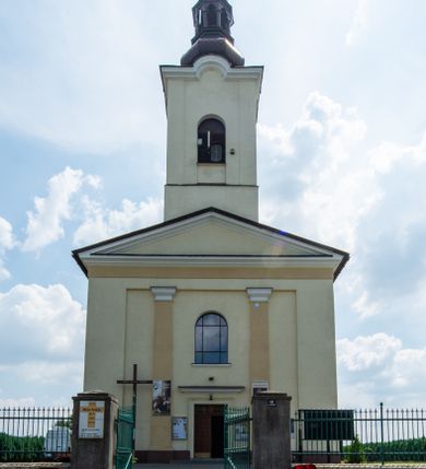 Zdjęcie nr 1: Kościół, zwrócony w kierunku południowo-wschodnim, jest budowlą murowaną z cegły na zaprawie wapiennej, na rzucie podłużnym. Powierzchnia budynku wynosi około 550 m2. Kościół charakteryzuje zwarta, prosta bryła. Korpus jest jednonawowy i trójprzęsłowy. Szerokie, dwuprzęsłowe, prezbiterium jest zamknięte półkoliście. Na osi kościoła, w przedłużeniu północnego przęsła przylega jednokondygnacyjna wieża, ujęta po bokach piętrowymi lokalnościami. Nawa przekryta jest sklepieniem kolebkowym z gurtami, wspartymi na osadzonych na wysokich cokołach parach przyściennych, czworobocznych filarów z przylegającymi do nich pilastrami o gładkich trzonach. Przestrzeń prezbiterium od korpusu nawowego oddziela półkoliście zamknięta arkada łuku tęczowego i drewniana balustrada. Zamknięte prostą ścianą prezbiterium zajmuje przestrzeń jednego przęsła. Dalej znajduje się zakrystia. Artykulację ścian na zewnątrz kościoła stanowi niewysoki obiegający bryłę cokół oraz niepełne belkowanie wieńczące, składające się z fryzu i gzymsu. Fasadę artykułuje płycina podzielona parą wspartych na cokołach pilastrów. W polu środkowym znajduje się główny otwór wejściowy. Dwuskrzydłowe, drewniane drzwi z naświetlem ujęte są prostym, profilowanym portalem. Tuż nad nadprożem znajduje się krótki gzyms. Wyżej duże, prostokątne, zamknięte półkoliście okno. Fasadę wieńczy trójkątny przyczółek. Ściany korpusu nawowego i prezbiterium przeprute są prostokątnymi, zamkniętymi półkoliście oknami. Dodatkowo w ścianie zachodniej, na wysokości przyległości wieży, w dolnej partii muru znajduje się niewielkie, półkoliście zamknięte okno a w drugim przęśle nawy prostokątny otwór drzwiowy; podobny jest umiejscowiony w drugim przęśle prezbiterium po stronie zachodniej. Kondygnację czworobocznej wieży w dolnej partii obiega parapet, wyżej w każdej ze ścian znajduje się po jednym prostokątnym, zamkniętym półkoliście oknie. Górną partię wieży obiega gzyms wieńczący wyłamujący się półkoliście pośrodku każdej ze ścian. Wieża nakryta jest hełmem cebulastym z latarnią. Korpus nawowy i prezbiterium przekrywa dwuspadowy dach wykonany z blachy, z wieżyczką na sygnaturkę na zakończeniu prezbiterium. Kolorystyka tynków kościoła utrzymana jest w odcieniach żółci i bieli, cokół jest brązowy.  Okna nawy, prezbiterium i fasady wypełniają witraże, wnętrze pokrywa polichromia z przedstawieniami figuralnymi w partii sklepień. Plac przykościelny, z wybrukowanymi alejkami i trawnikiem, otoczony jest żywopłotem z krzewów ozdobnych.