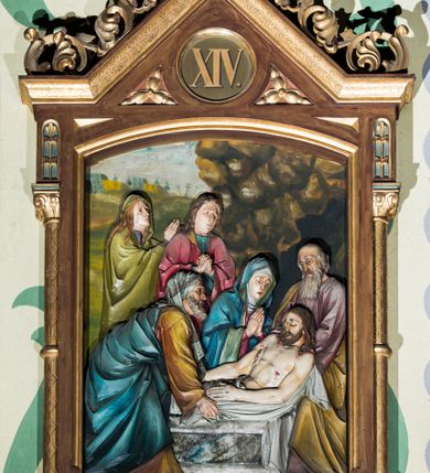 Zdjęcie nr 13: Czternaście płaskorzeźbionych stacji Drogi krzyżowej składa się z przedstawienia ujętego w taką samą architektoniczną, drewnianą ramę. Pole obrazowe uzyskał kształt stojącego prostokąta zamkniętego u góry łukiem odcinkowym. Flankują go smukłe półkolumienki wsparte na niskich bazach, ustawionych na prostokątnym, profilowanym cokole wspartym od dołu po bokach niewielkimi konsolami oraz udekorowanym podwieszonym na osi stylizowanym ornamentem roślinnym. Na cokole umieszczono podpis poszczególnej stacji. Kolumienki podtrzymują profilowane belkowanie w kształcie nadwieszonego trójkąta zwieńczonego krzyżem. Zewnętrze boki trójkąta zdobi ażurowy, złocony ornament roślinny. W polu trójkąta w medalionie wpisano kolejny numer stacji. Strukturę obramienia polichromowano na brązowo, natomiast kolumienki, wieńczące krzyże, ornamenty oraz profile zostały pozłocone. W polach obrazowych znalazły się wielopostaciowe, płaskorzeźbione kompozycje umieszczone najczęściej na tle pejzażu i architektury. Narracja każdego z obrazów skupia się na osobie Chrystusa, któremu towarzyszą inni ludzie, zgodnie z narracją kolejnej stacji. Przedstawione postacie wykonują gwałtowne i dynamiczne gesty. Na ich twarzach o jasnych karnacjach widoczne są różne emocje. Postacie ubrane są w obficie drapowane szaty o intensywnej i kontrastowej kolorystyce. Cykl przedstawia stacje z następującymi podpisami: I. Pan Jezus przez Piłata na śmierć skazany, II. Pan Jezus bierze krzyż na barki, III. Pan Jezus upada po raz pierwszy, IV. Pan Jezus spotyka się ze swoją Matką, V. Panu Jezusowi dopomaga Szymon Cyrenejczyk, VI. Panu Jezusowi ociera twarz św. Weronika, VII. Pan Jezus upada drugi raz, VIII. Pan Jezus pociesza płaczące niewiasty, IX. Pan Jezus upada po raz trzeci, X. Pan Jezus obnażony i żółcią napawany, XI. Pan Jezus do krzyża przybijany, XII. Pan Jezus umiera na krzyżu, XIII. Pan Jezus z krzyża zdjęty, XIV. Pan Jezus złożony w grobie. 