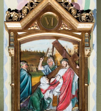 Zdjęcie nr 6: Czternaście płaskorzeźbionych stacji Drogi krzyżowej składa się z przedstawienia ujętego w taką samą architektoniczną, drewnianą ramę. Pole obrazowe uzyskał kształt stojącego prostokąta zamkniętego u góry łukiem odcinkowym. Flankują go smukłe półkolumienki wsparte na niskich bazach, ustawionych na prostokątnym, profilowanym cokole wspartym od dołu po bokach niewielkimi konsolami oraz udekorowanym podwieszonym na osi stylizowanym ornamentem roślinnym. Na cokole umieszczono podpis poszczególnej stacji. Kolumienki podtrzymują profilowane belkowanie w kształcie nadwieszonego trójkąta zwieńczonego krzyżem. Zewnętrze boki trójkąta zdobi ażurowy, złocony ornament roślinny. W polu trójkąta w medalionie wpisano kolejny numer stacji. Strukturę obramienia polichromowano na brązowo, natomiast kolumienki, wieńczące krzyże, ornamenty oraz profile zostały pozłocone. W polach obrazowych znalazły się wielopostaciowe, płaskorzeźbione kompozycje umieszczone najczęściej na tle pejzażu i architektury. Narracja każdego z obrazów skupia się na osobie Chrystusa, któremu towarzyszą inni ludzie, zgodnie z narracją kolejnej stacji. Przedstawione postacie wykonują gwałtowne i dynamiczne gesty. Na ich twarzach o jasnych karnacjach widoczne są różne emocje. Postacie ubrane są w obficie drapowane szaty o intensywnej i kontrastowej kolorystyce. Cykl przedstawia stacje z następującymi podpisami: I. Pan Jezus przez Piłata na śmierć skazany, II. Pan Jezus bierze krzyż na barki, III. Pan Jezus upada po raz pierwszy, IV. Pan Jezus spotyka się ze swoją Matką, V. Panu Jezusowi dopomaga Szymon Cyrenejczyk, VI. Panu Jezusowi ociera twarz św. Weronika, VII. Pan Jezus upada drugi raz, VIII. Pan Jezus pociesza płaczące niewiasty, IX. Pan Jezus upada po raz trzeci, X. Pan Jezus obnażony i żółcią napawany, XI. Pan Jezus do krzyża przybijany, XII. Pan Jezus umiera na krzyżu, XIII. Pan Jezus z krzyża zdjęty, XIV. Pan Jezus złożony w grobie. 