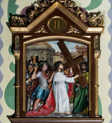 Zdjęcie nr 2: Czternaście płaskorzeźbionych stacji Drogi krzyżowej składa się z przedstawienia ujętego w taką samą architektoniczną, drewnianą ramę. Pole obrazowe uzyskał kształt stojącego prostokąta zamkniętego u góry łukiem odcinkowym. Flankują go smukłe półkolumienki wsparte na niskich bazach, ustawionych na prostokątnym, profilowanym cokole wspartym od dołu po bokach niewielkimi konsolami oraz udekorowanym podwieszonym na osi stylizowanym ornamentem roślinnym. Na cokole umieszczono podpis poszczególnej stacji. Kolumienki podtrzymują profilowane belkowanie w kształcie nadwieszonego trójkąta zwieńczonego krzyżem. Zewnętrze boki trójkąta zdobi ażurowy, złocony ornament roślinny. W polu trójkąta w medalionie wpisano kolejny numer stacji. Strukturę obramienia polichromowano na brązowo, natomiast kolumienki, wieńczące krzyże, ornamenty oraz profile zostały pozłocone. W polach obrazowych znalazły się wielopostaciowe, płaskorzeźbione kompozycje umieszczone najczęściej na tle pejzażu i architektury. Narracja każdego z obrazów skupia się na osobie Chrystusa, któremu towarzyszą inni ludzie, zgodnie z narracją kolejnej stacji. Przedstawione postacie wykonują gwałtowne i dynamiczne gesty. Na ich twarzach o jasnych karnacjach widoczne są różne emocje. Postacie ubrane są w obficie drapowane szaty o intensywnej i kontrastowej kolorystyce. Cykl przedstawia stacje z następującymi podpisami: I. Pan Jezus przez Piłata na śmierć skazany, II. Pan Jezus bierze krzyż na barki, III. Pan Jezus upada po raz pierwszy, IV. Pan Jezus spotyka się ze swoją Matką, V. Panu Jezusowi dopomaga Szymon Cyrenejczyk, VI. Panu Jezusowi ociera twarz św. Weronika, VII. Pan Jezus upada drugi raz, VIII. Pan Jezus pociesza płaczące niewiasty, IX. Pan Jezus upada po raz trzeci, X. Pan Jezus obnażony i żółcią napawany, XI. Pan Jezus do krzyża przybijany, XII. Pan Jezus umiera na krzyżu, XIII. Pan Jezus z krzyża zdjęty, XIV. Pan Jezus złożony w grobie. 